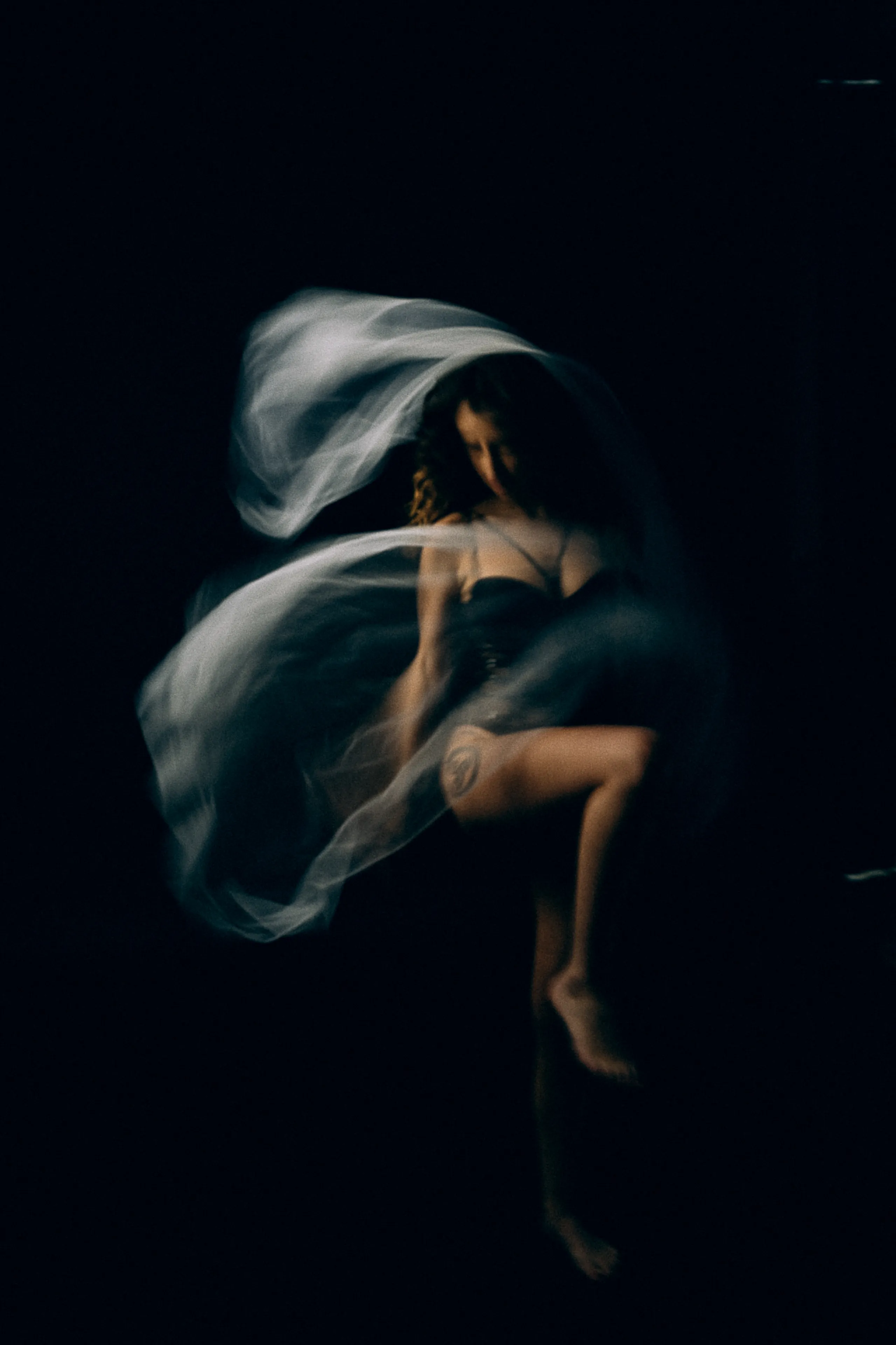 A figure in motion beneath flying sheer fabric, warm skin against black