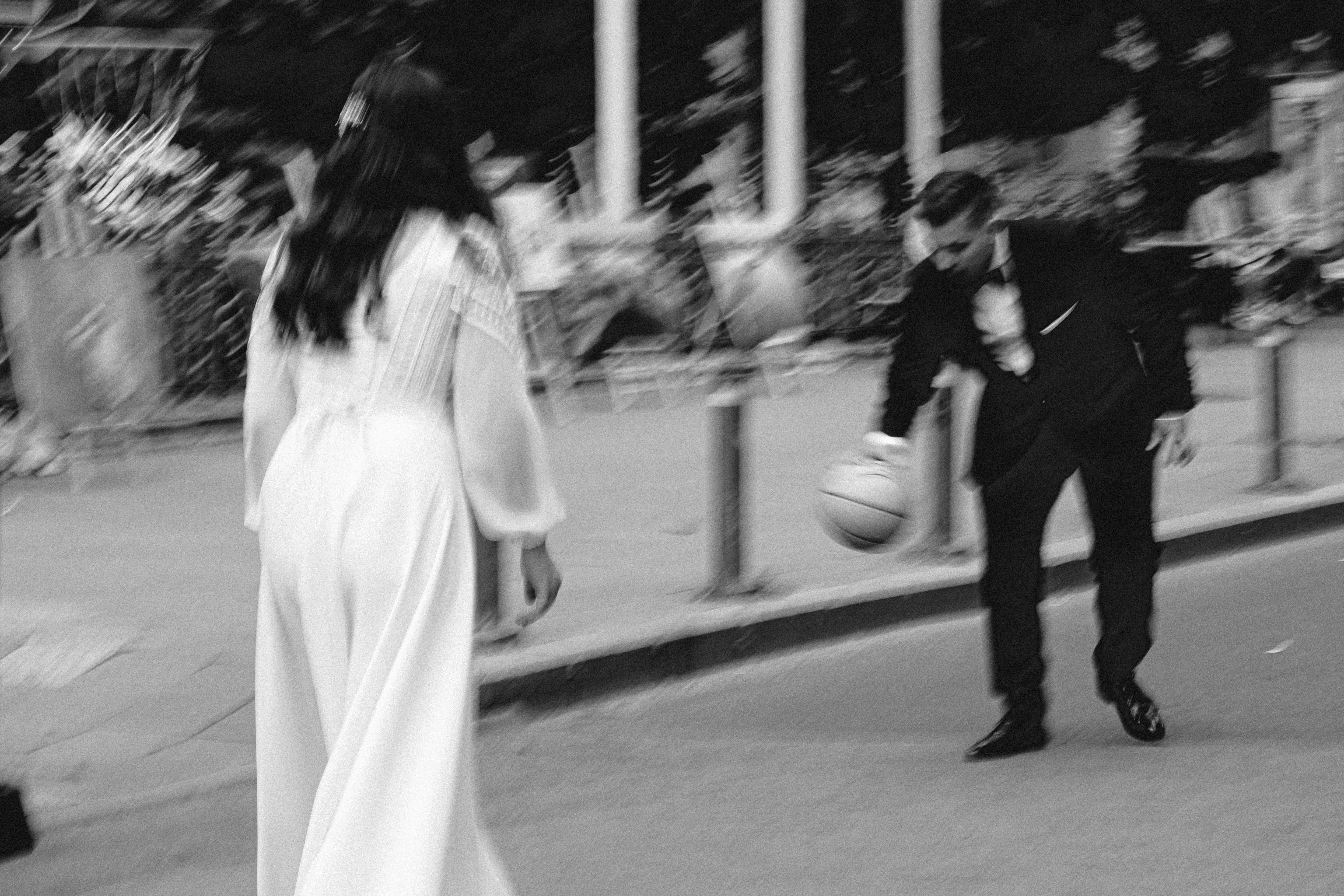 A bride and groom on a city street in motion blur — he dribbles a basketball on the curb while she walks toward him, both mid-laugh