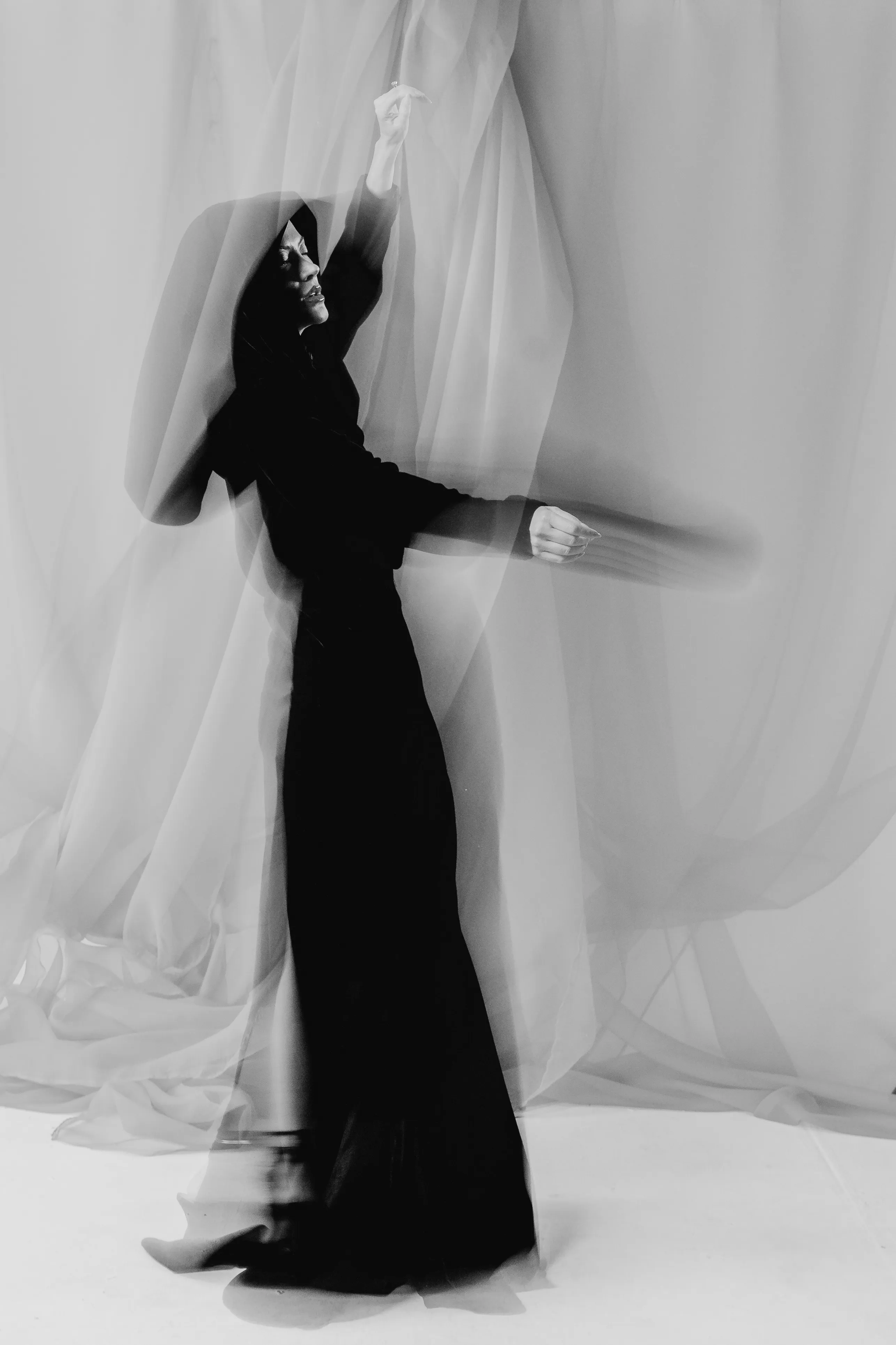 A woman in a black gown moves beneath sheer draped fabric, in black and white