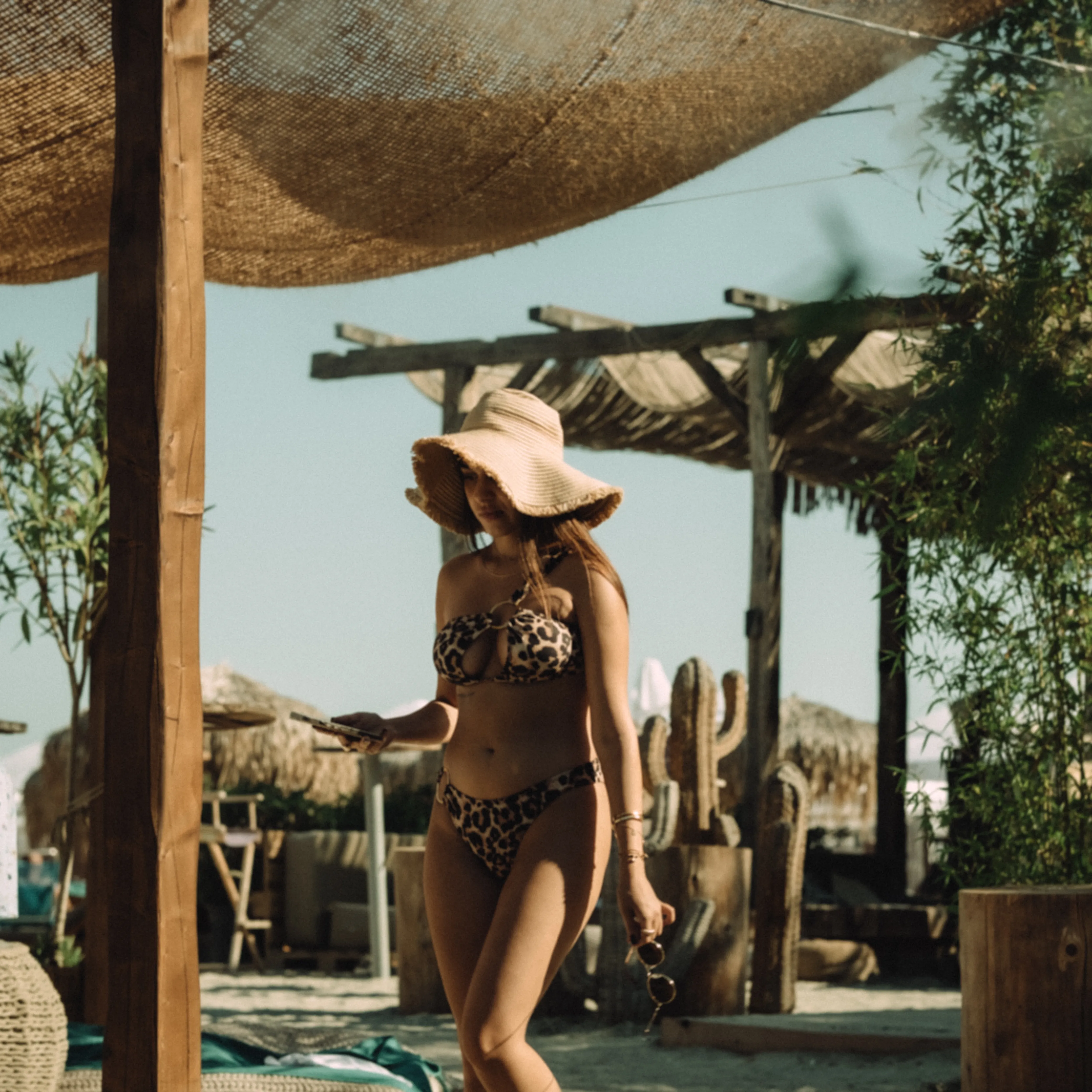 A woman in a leopard-print swimsuit walks under a wooden pergola in golden light