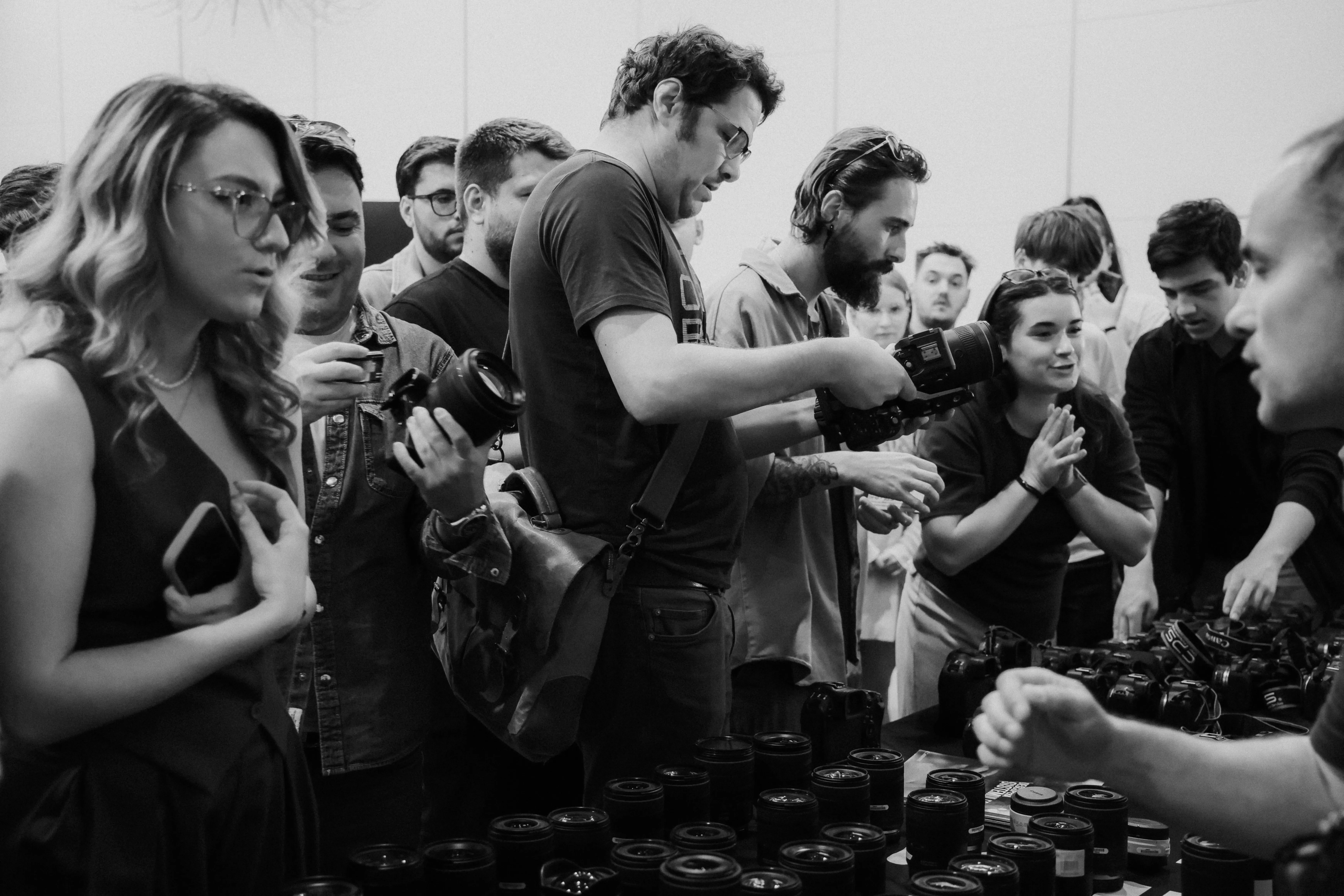 Black and white candid of a crowd gathered around a table of lenses, testing and comparing gear