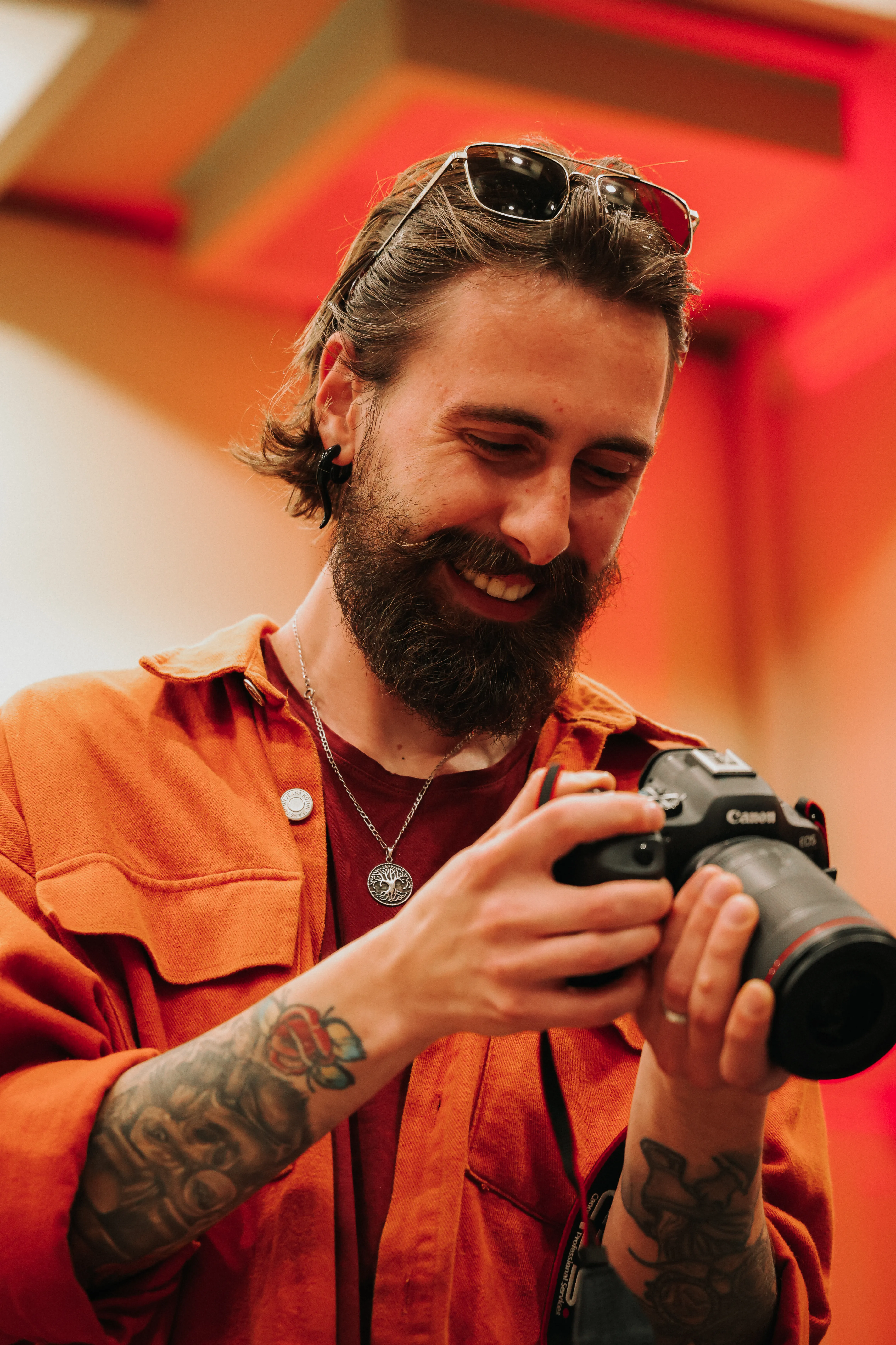 A man in an orange jacket and sunglasses smiles while holding a camera to his chest