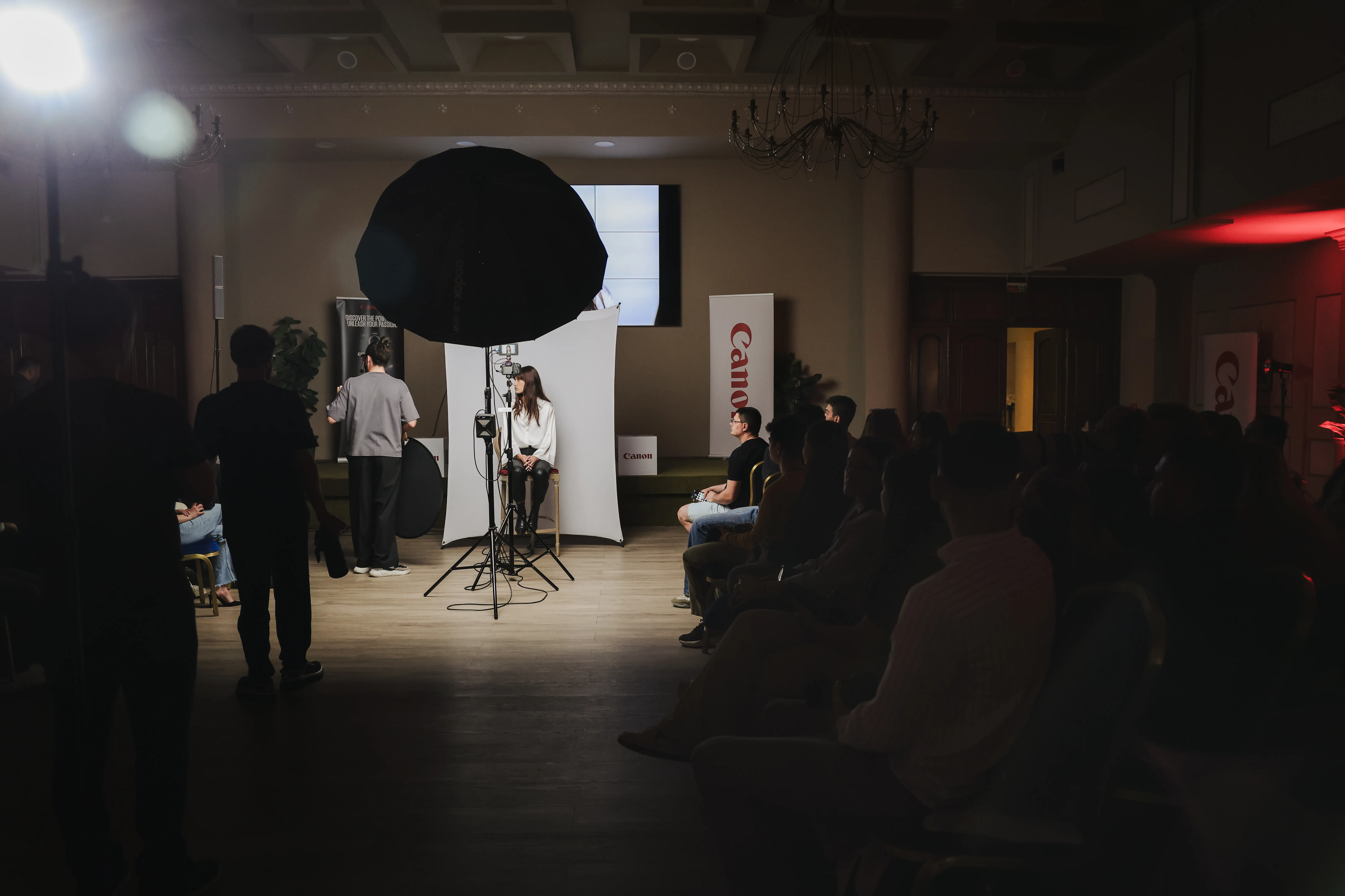 Wide view of a chandeliered ballroom set up for a Canon masterclass, a model lit under a softbox as the audience watches
