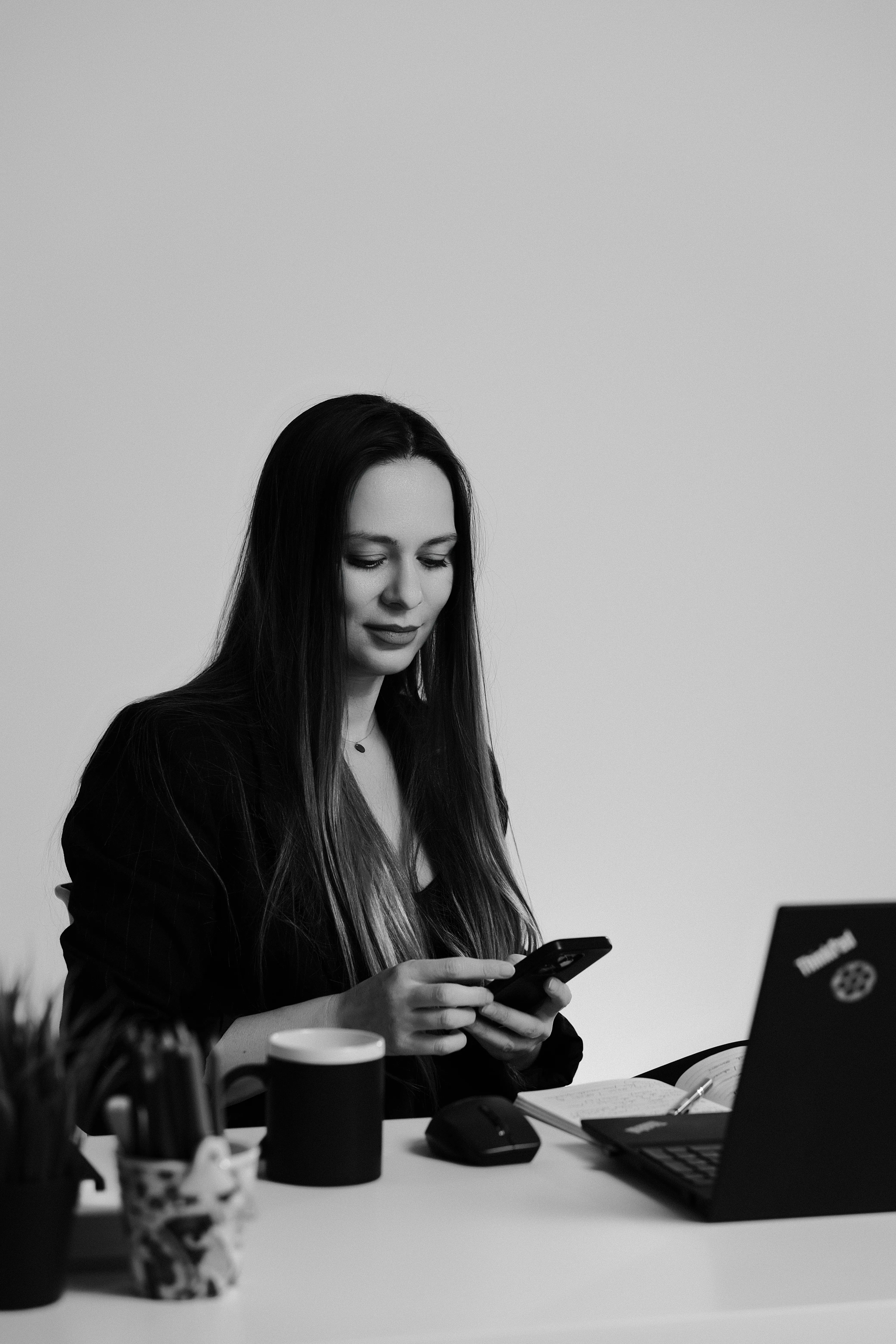 Black and white portrait of a woman at her desk, looking at her phone, laptop in frame