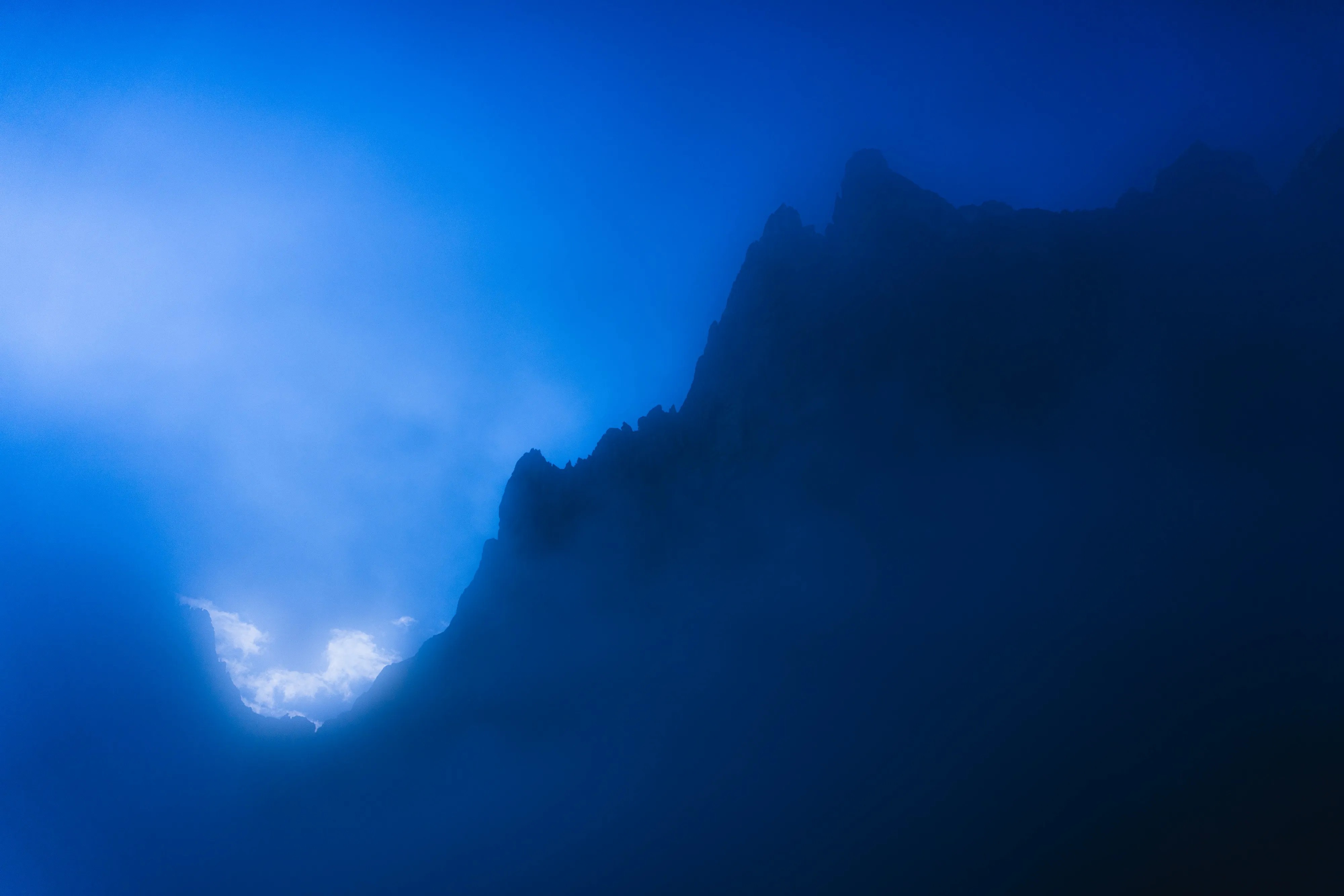 A mountain peak silhouetted in deep blue, a break of cloud catching the light beside it