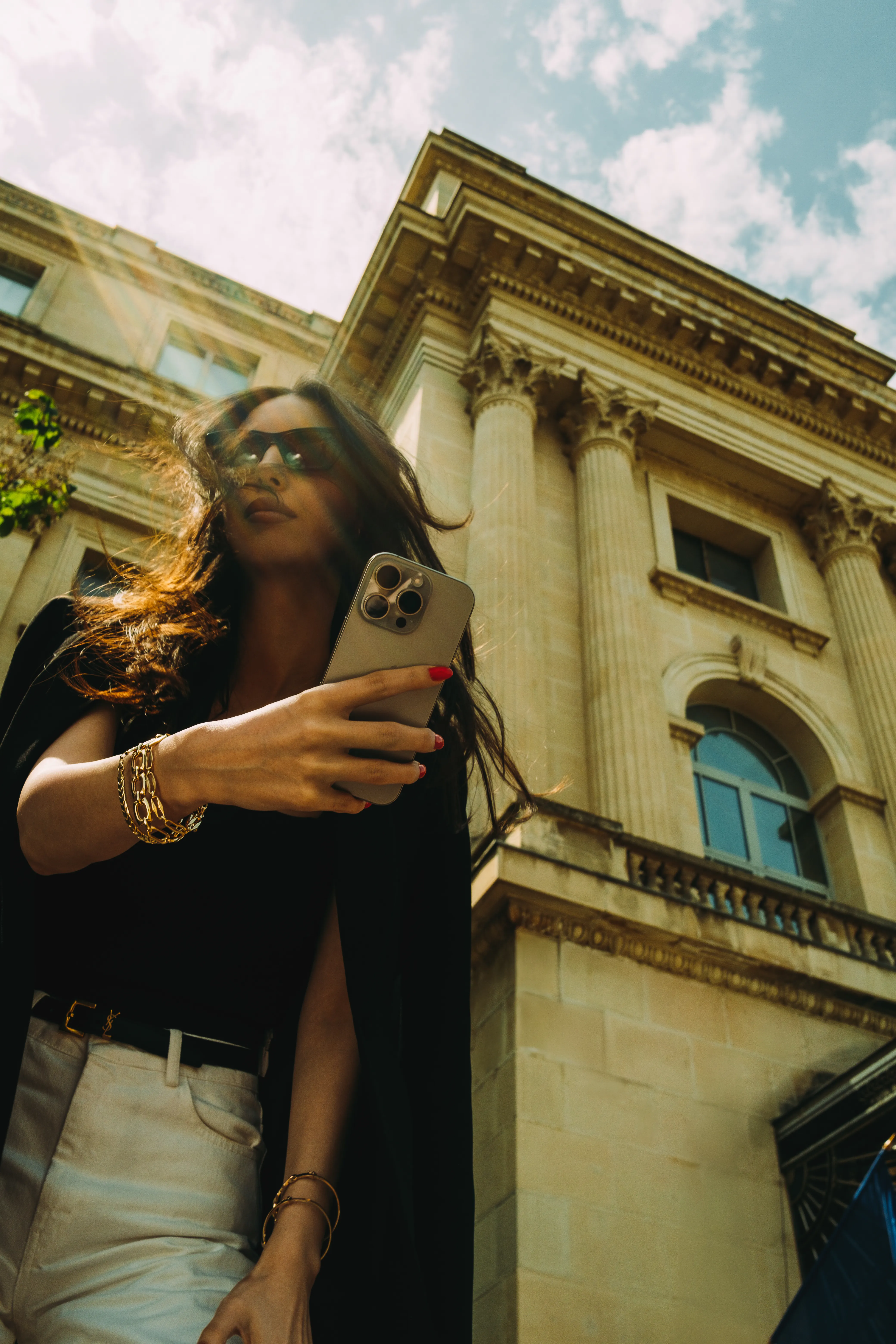 Low-angle shot of a woman in sunglasses holding a phone up, gold jewelry catching the light, Corinthian-columned building behind her