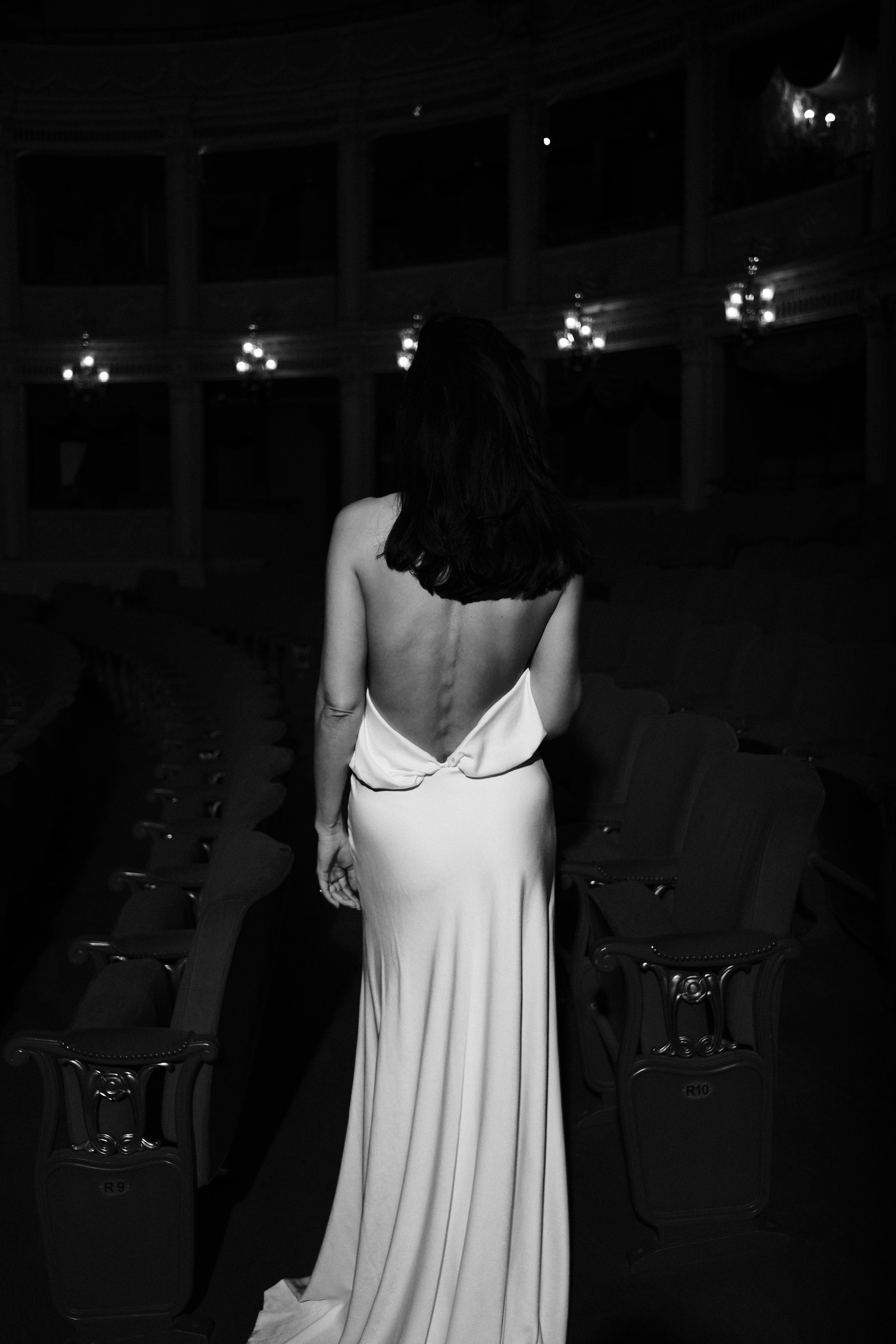 Woman reclined across a row of red velvet theatre seats in an evening dress, candlelit sconces along the wall