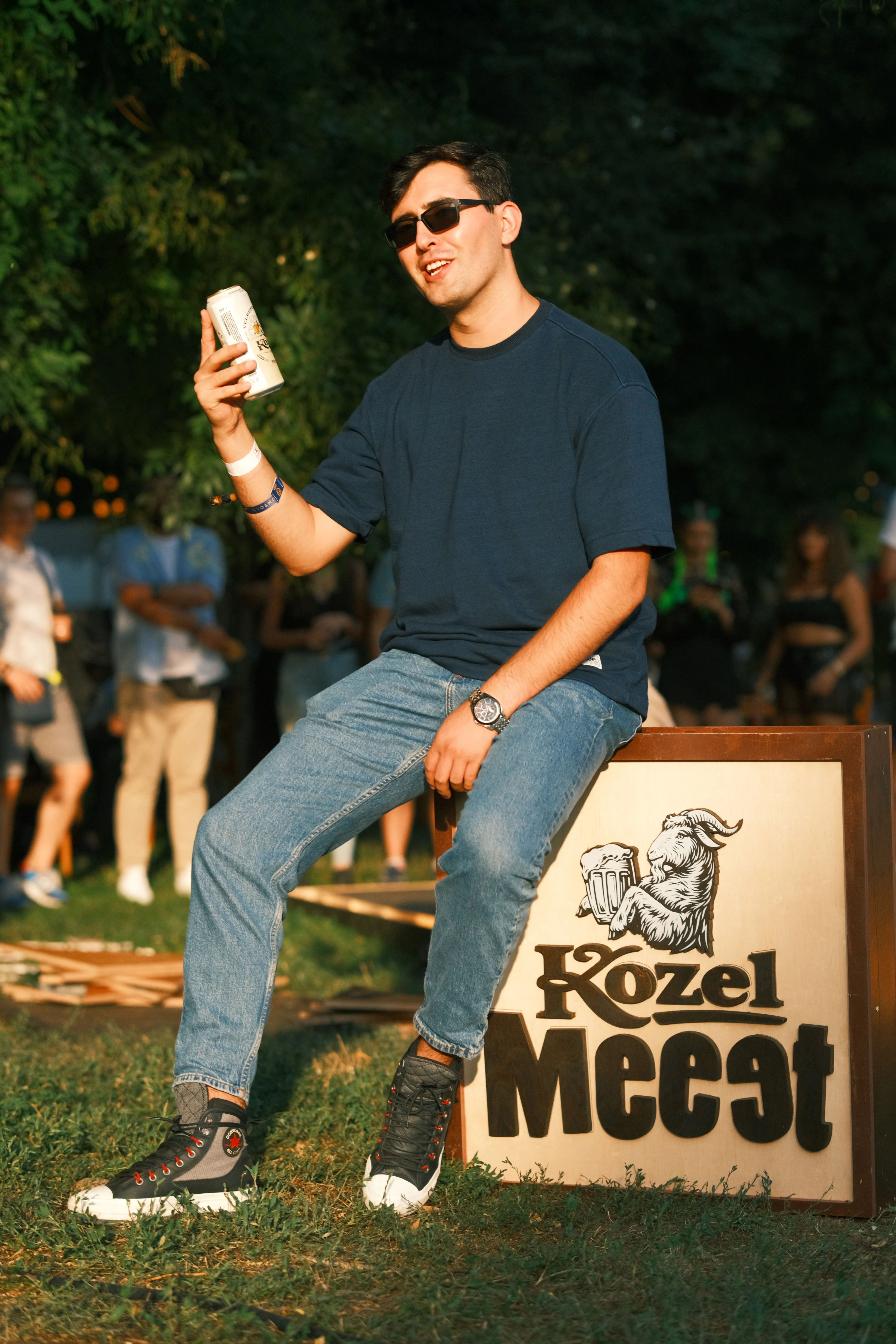 A man laughing and holding a can, sitting on a branded festival sign in evening light