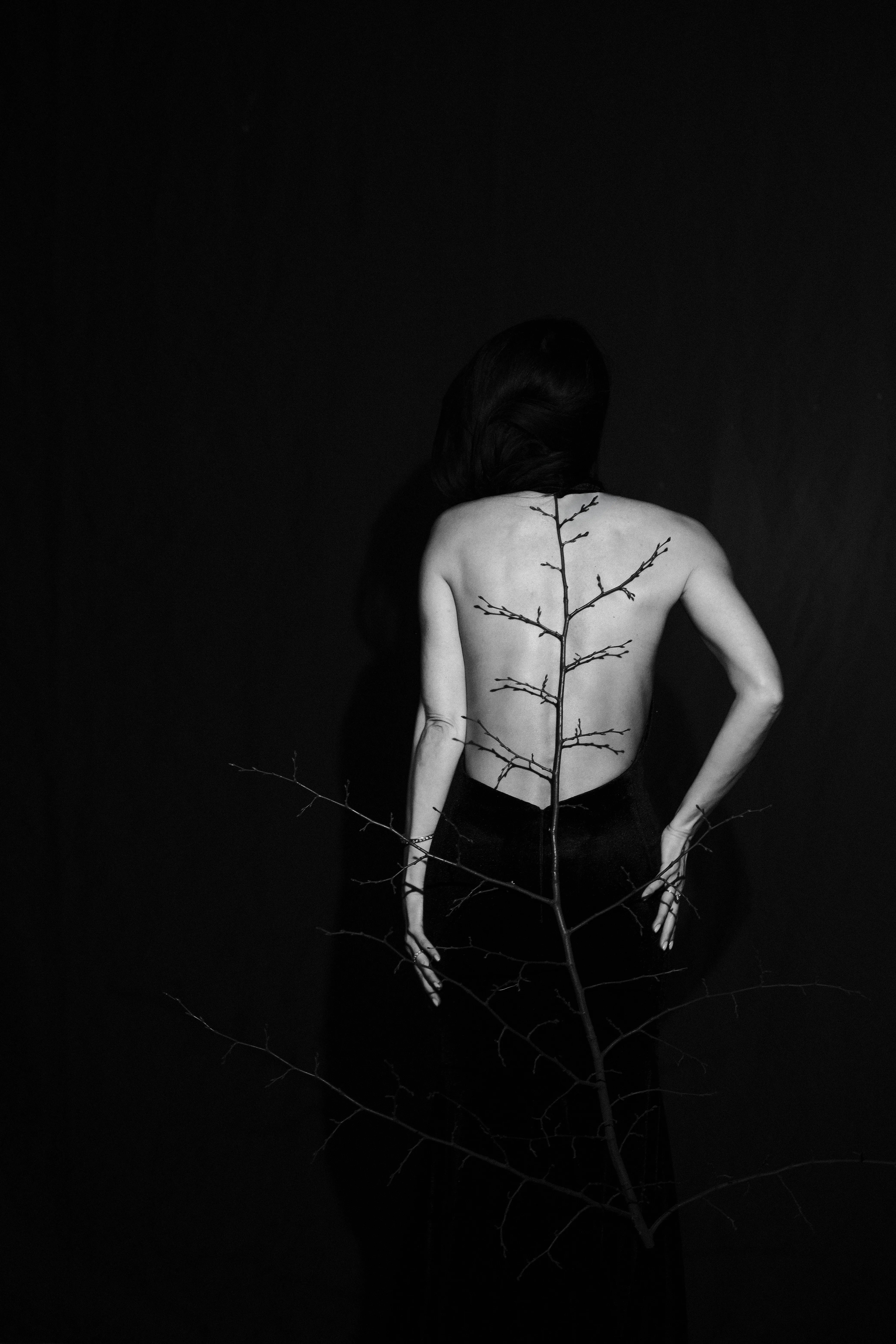 A standing figure from behind, a bare branch running the length of her spine, in black and white