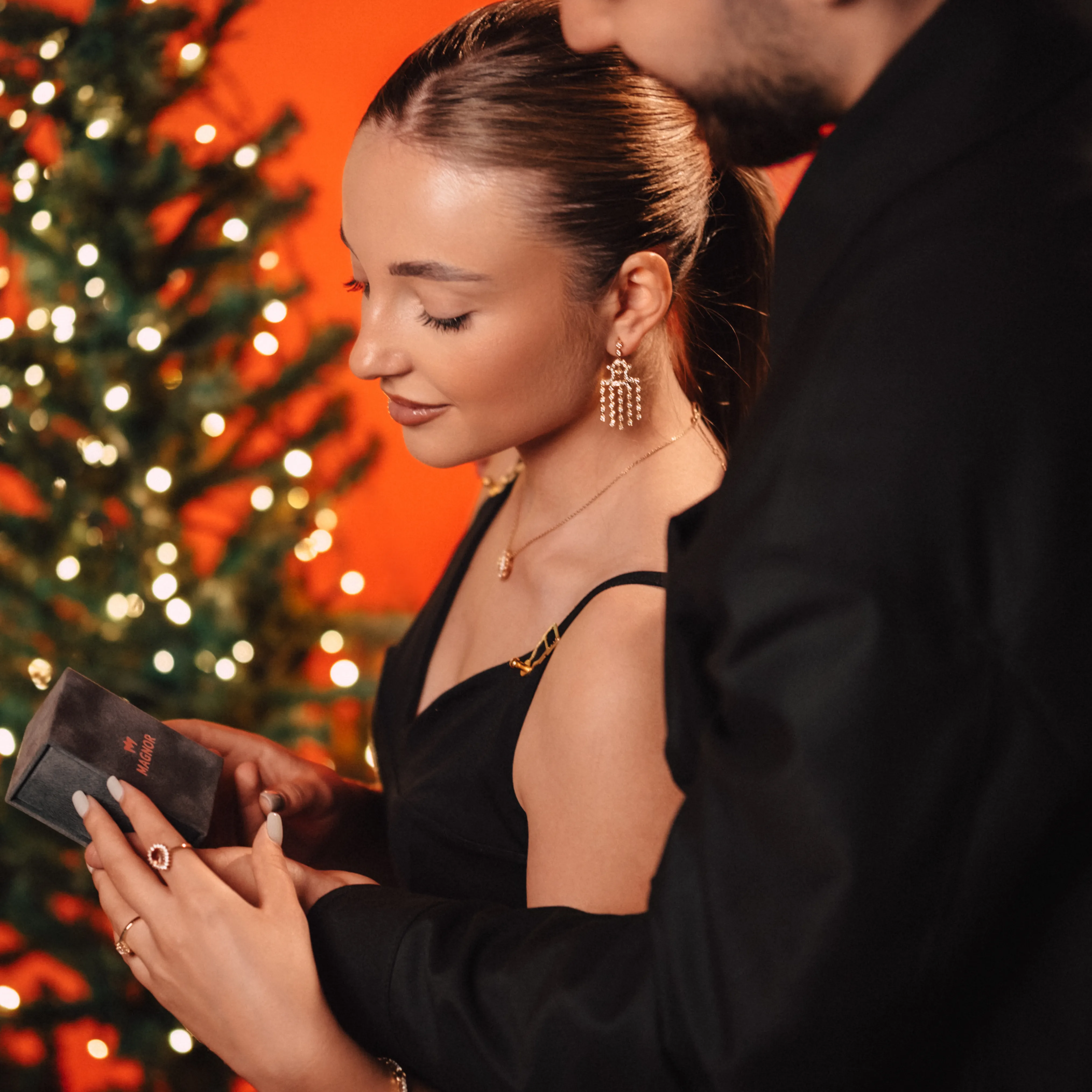 A couple stands close beside a Christmas tree strung with warm lights, jewelry on the woman's hand