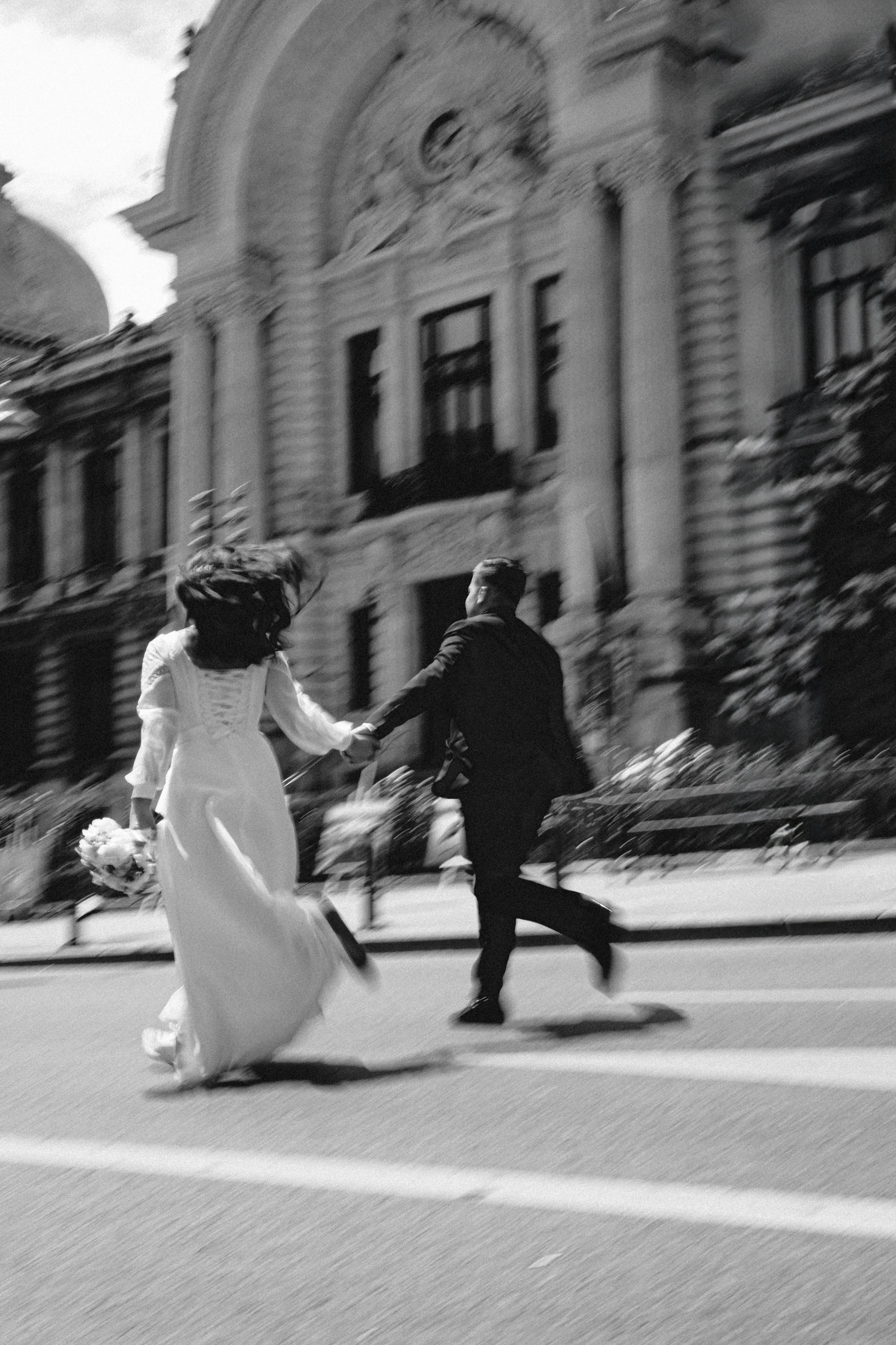 A couple in motion blur running hand in hand across a street, a grand ornate building behind them, black and white
