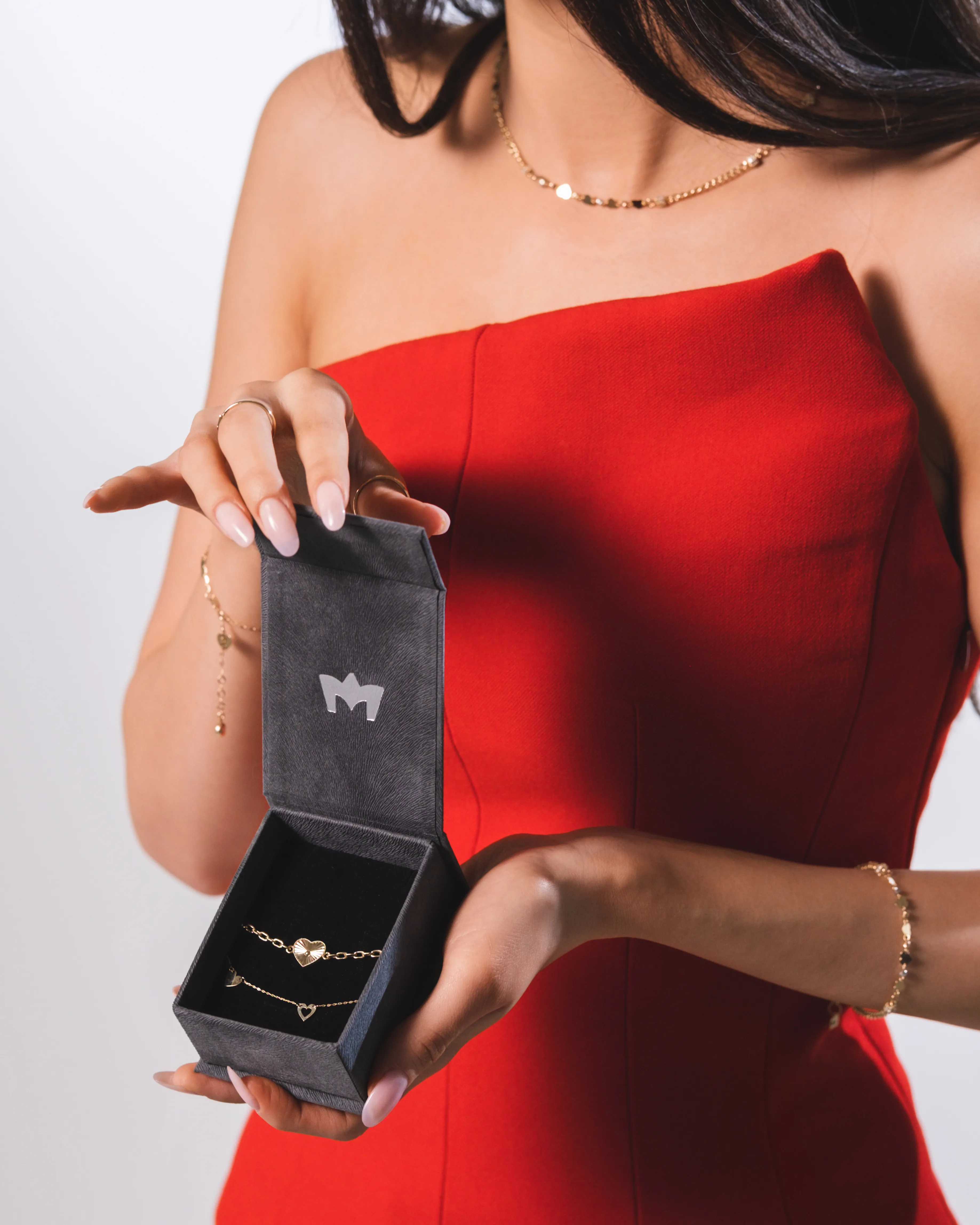 A woman in a red dress opens a jewelry gift box tied with ribbon, mid-laugh