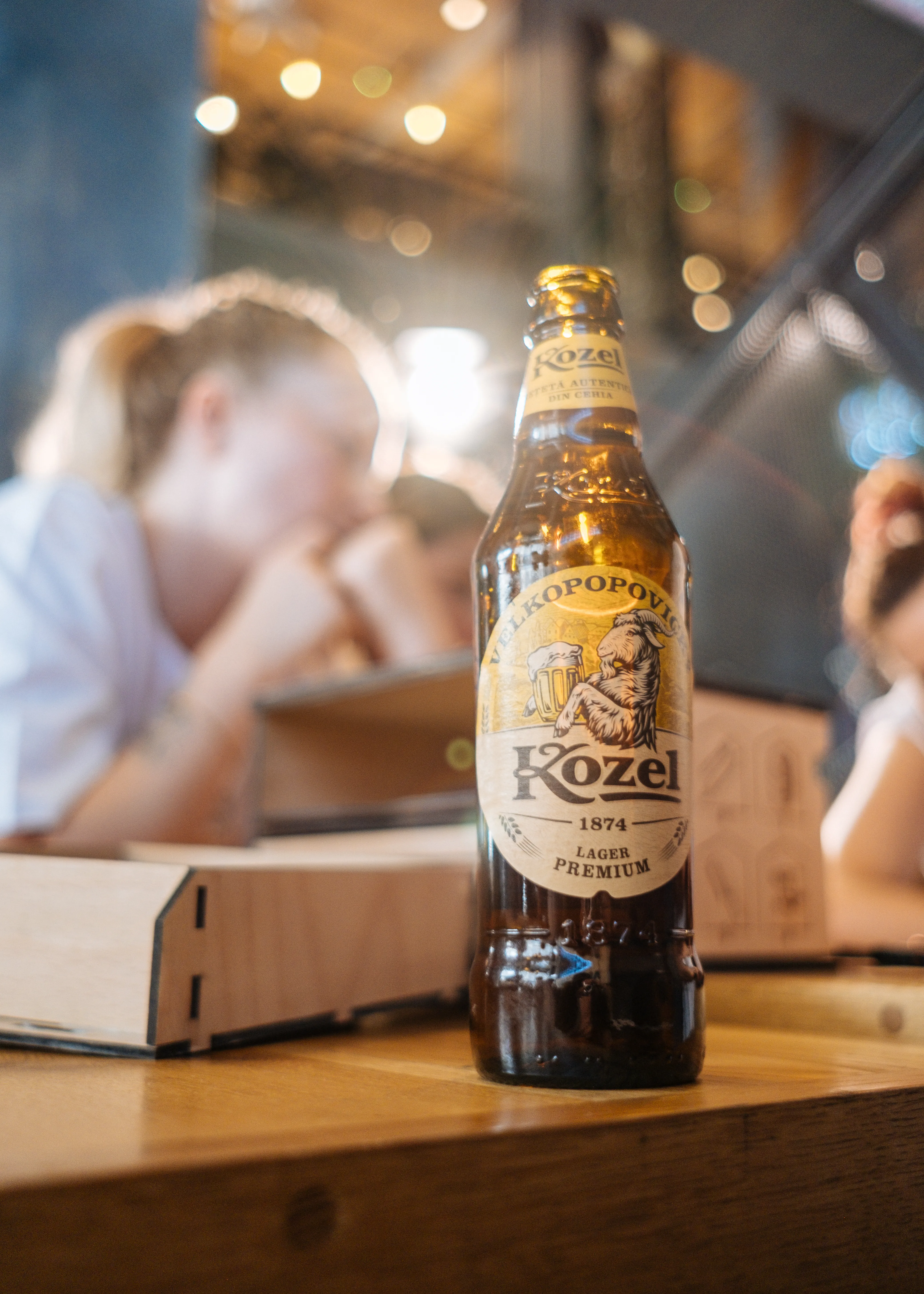 A Kozel beer bottle on a restaurant table, people talking softly out of focus behind it