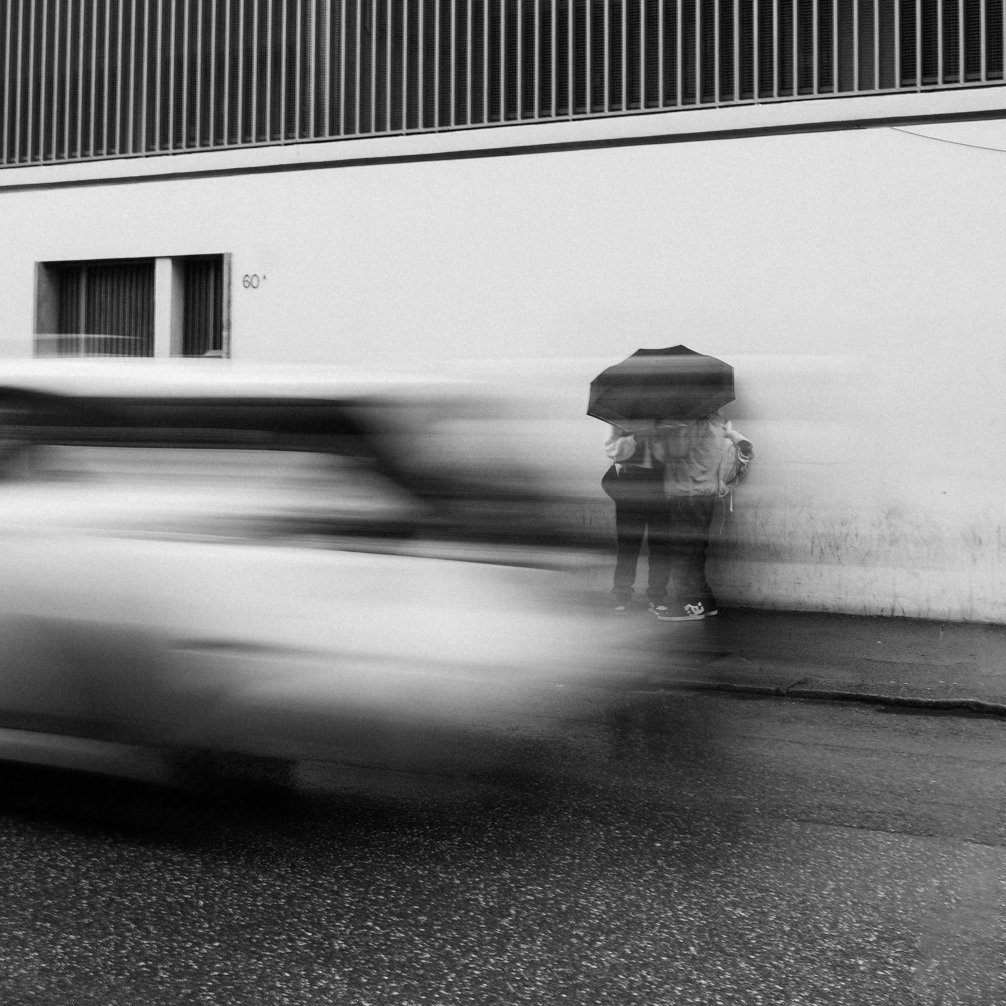 A couple sheltering under an umbrella against a wall as a car blurs past in the rain, black and white