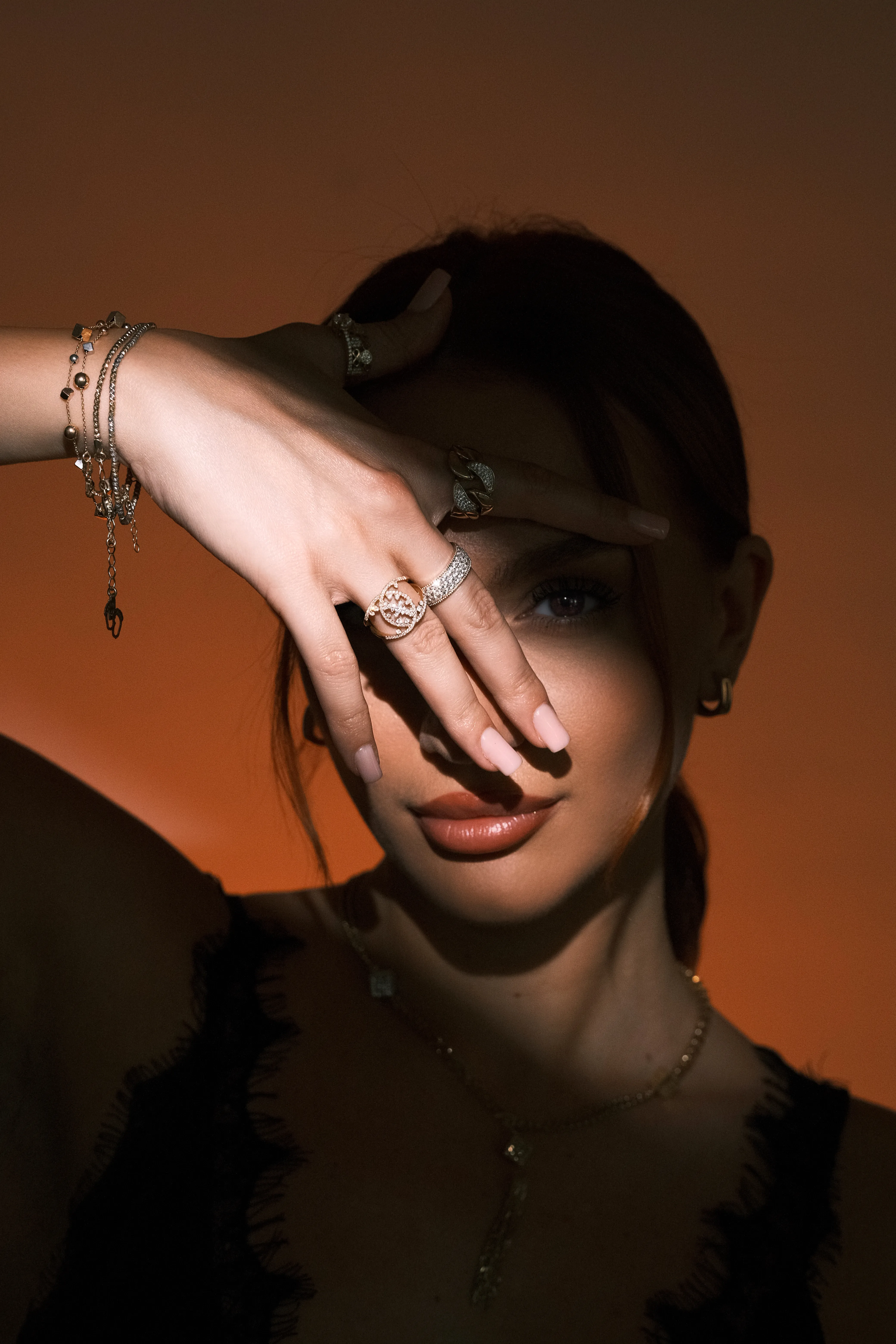 A dramatic hand-shield gesture against a warm gradient background, rings and bracelets lit hard