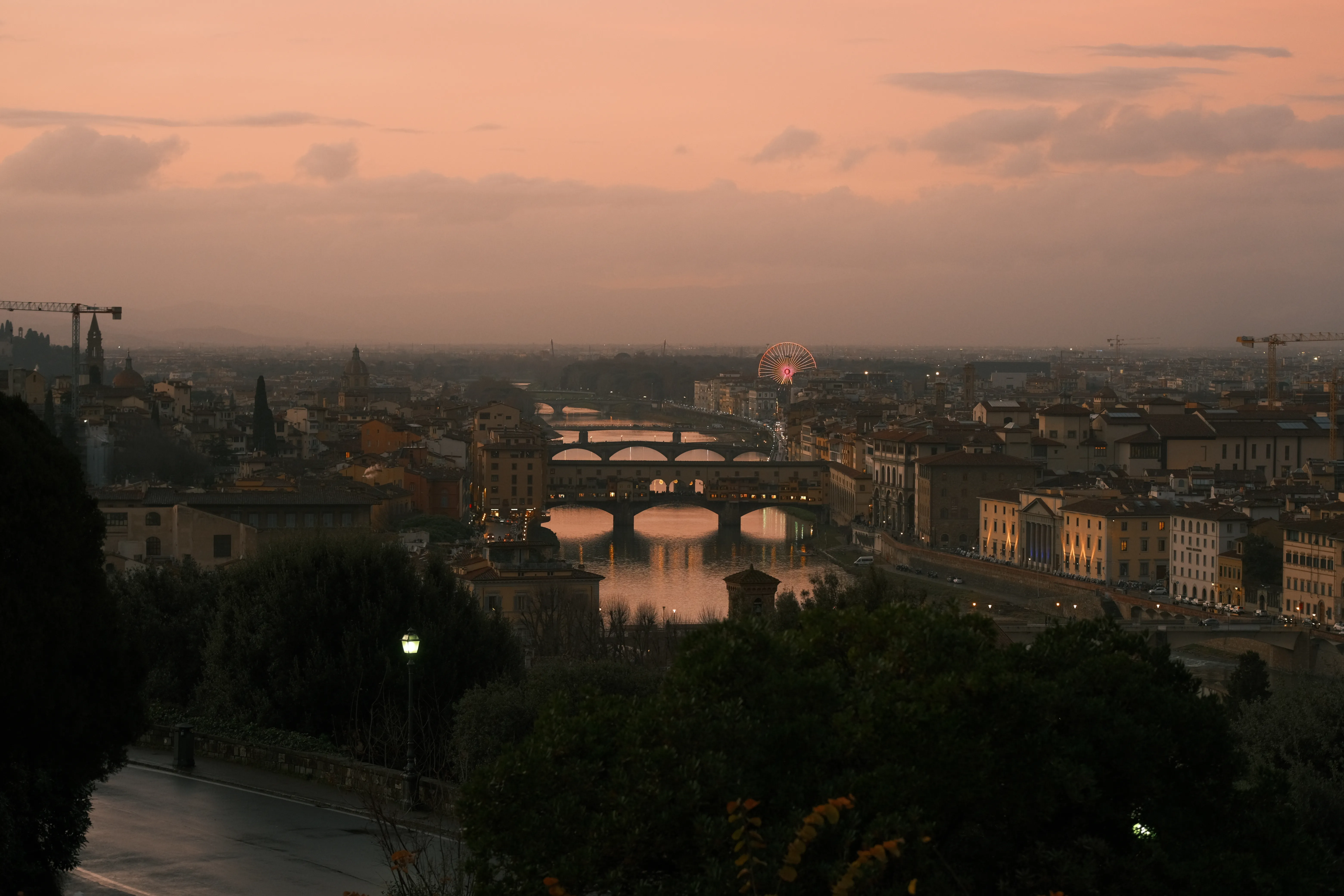 Ponte Vecchio and the Arno river seen from a hillside at dusk, warm pink sky, a small lit ferris wheel in the distance