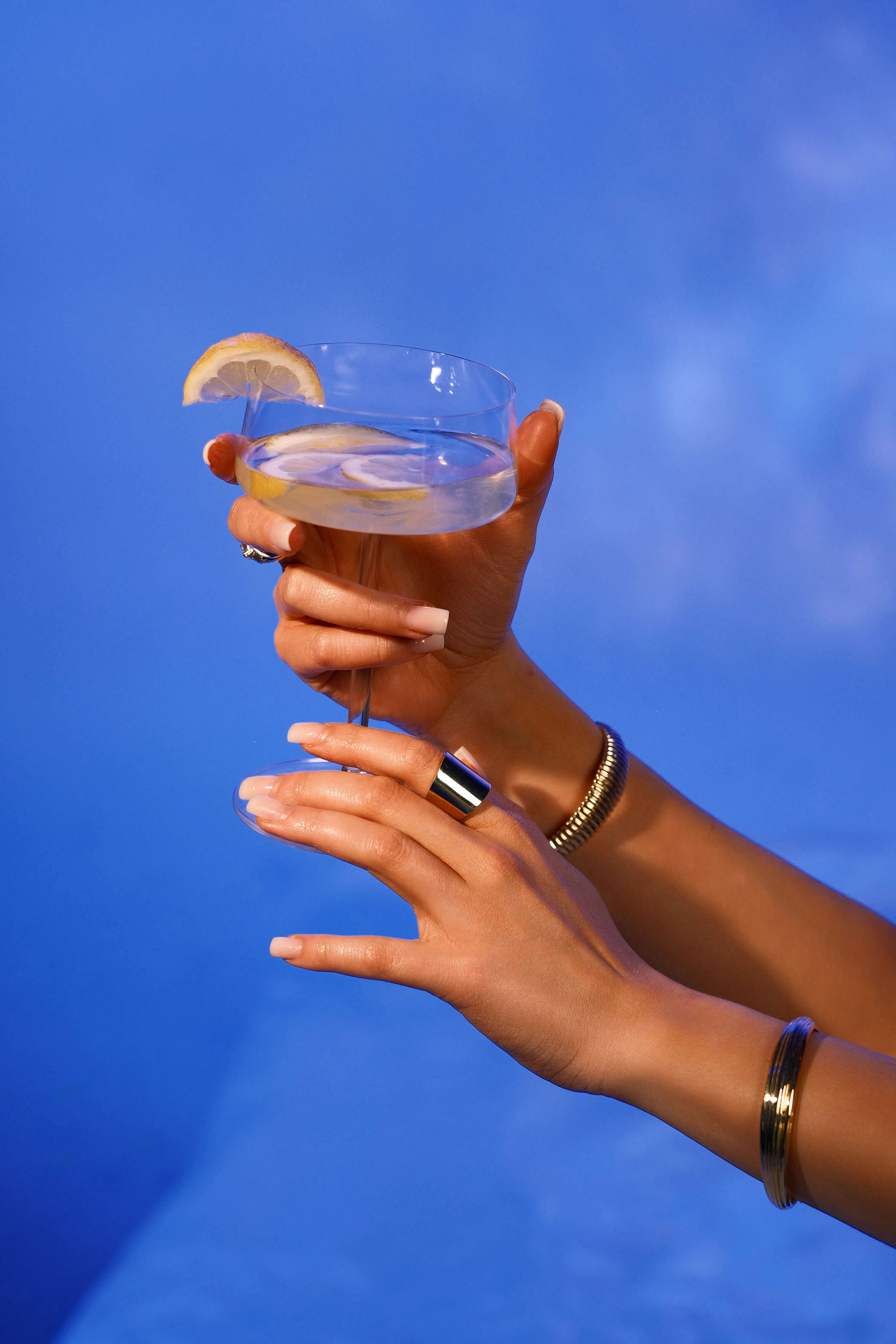 A hand wearing a ring holds a blue cocktail with a lemon garnish against a sky-blue background