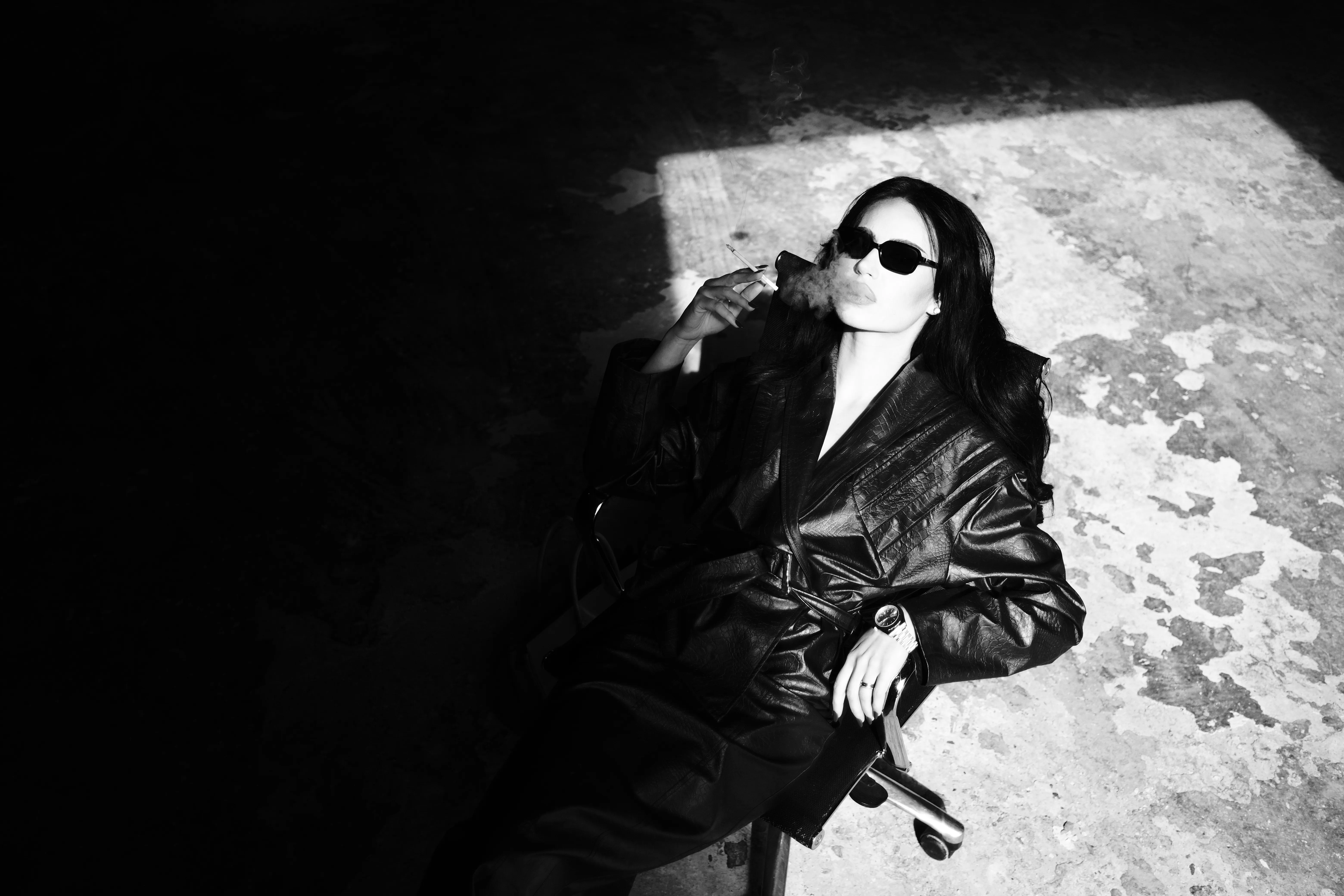 Woman in a leather coat and sunglasses smoking, dramatic overhead light, black and white