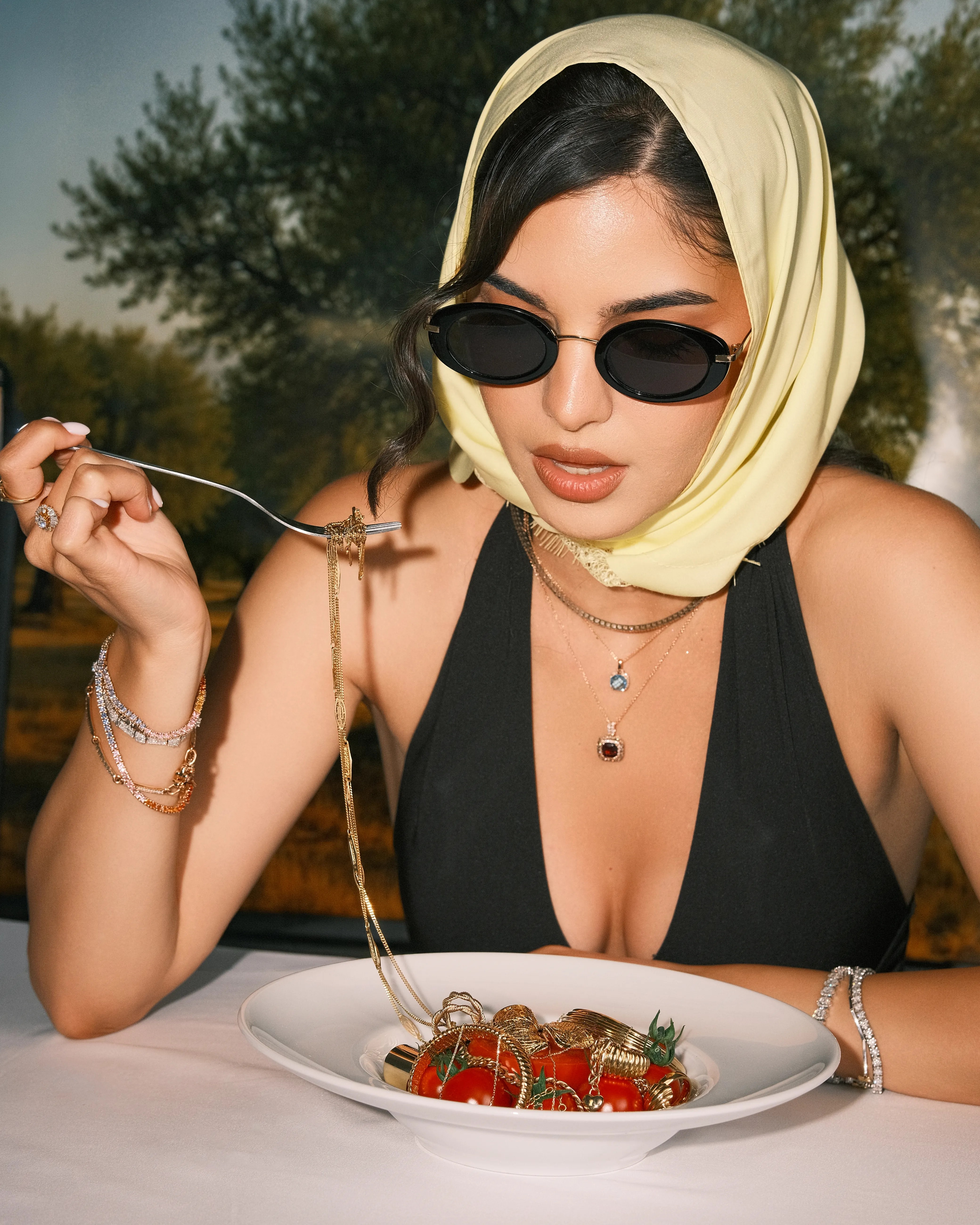 A woman in a headscarf and sunglasses twirls pasta at a sunlit outdoor table, jewelry on her hand