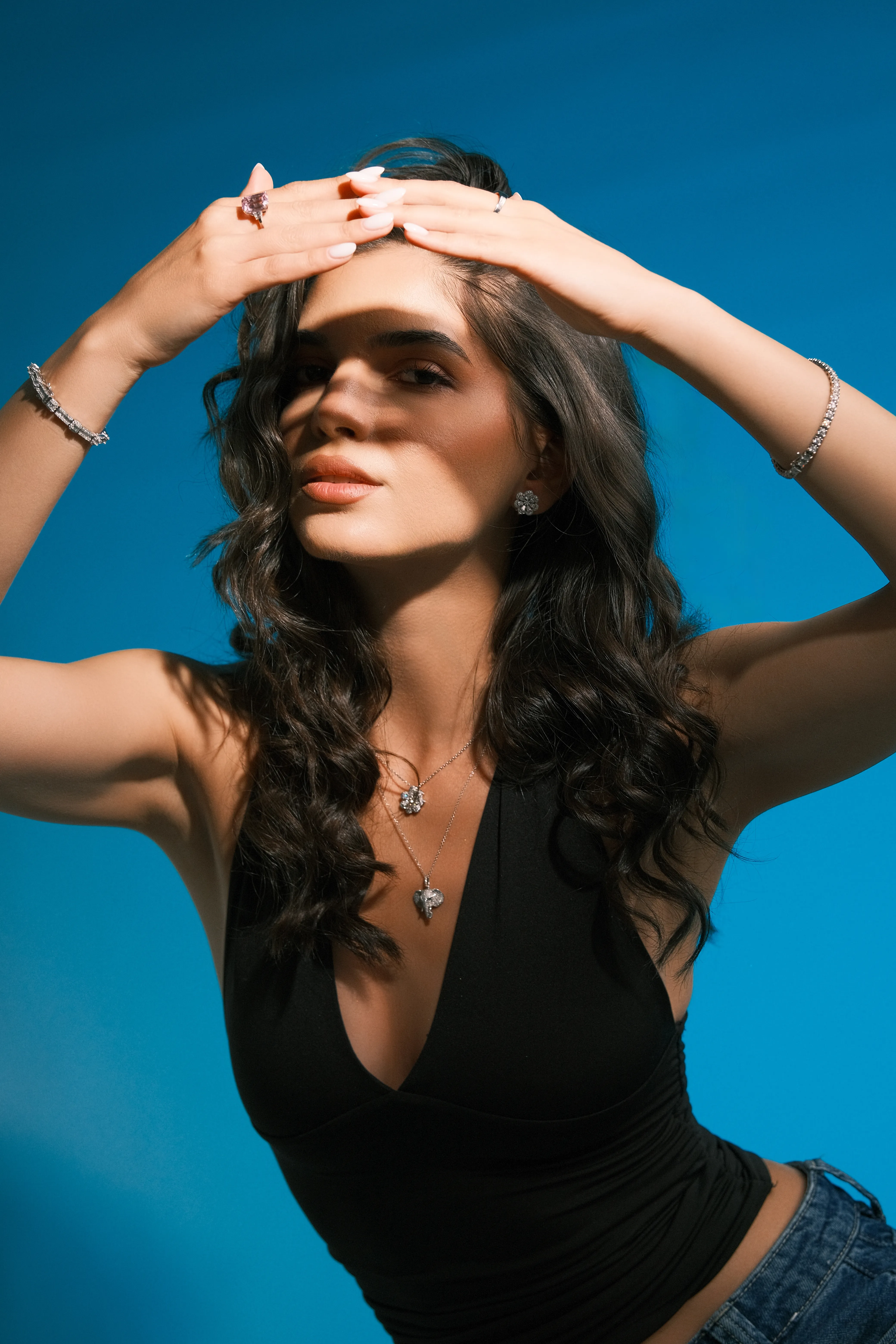 A woman in a black top raises both arms overhead, gold jewelry catching the light, against a sky-blue background