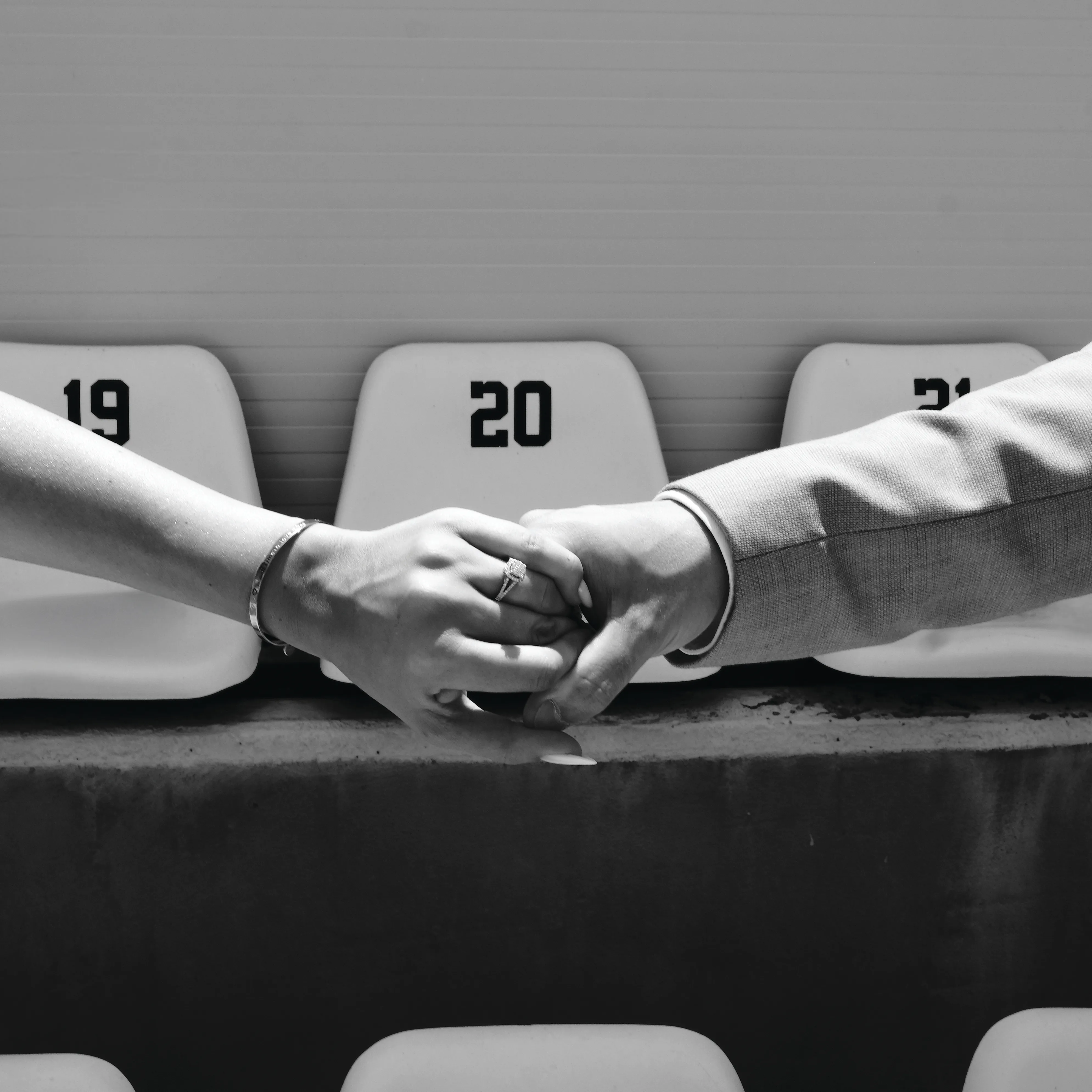 Two hands resting together on a stadium seat, wedding rings visible, black and white