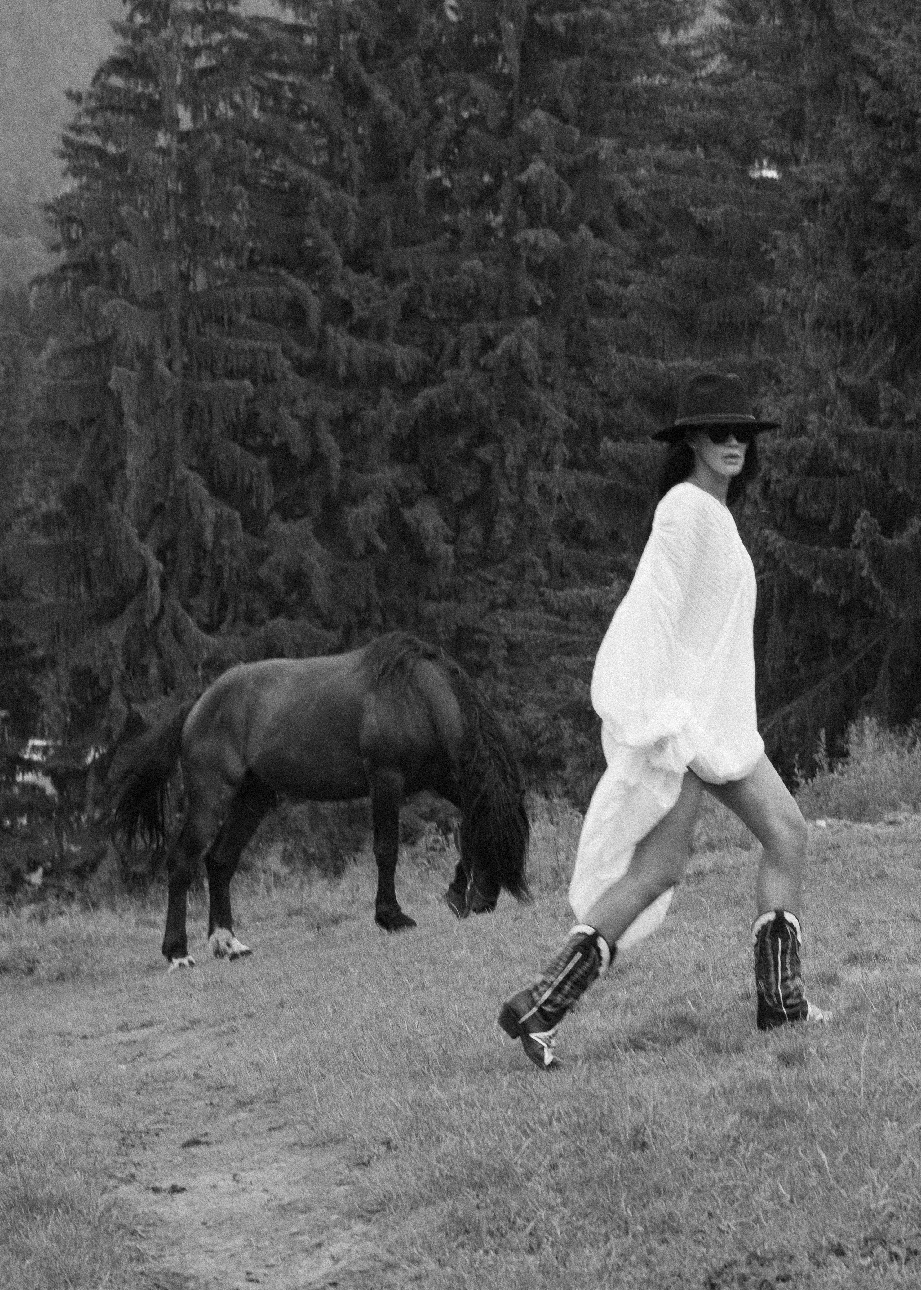 Black and white editorial portrait of a woman walking in a white dress beside a horse
