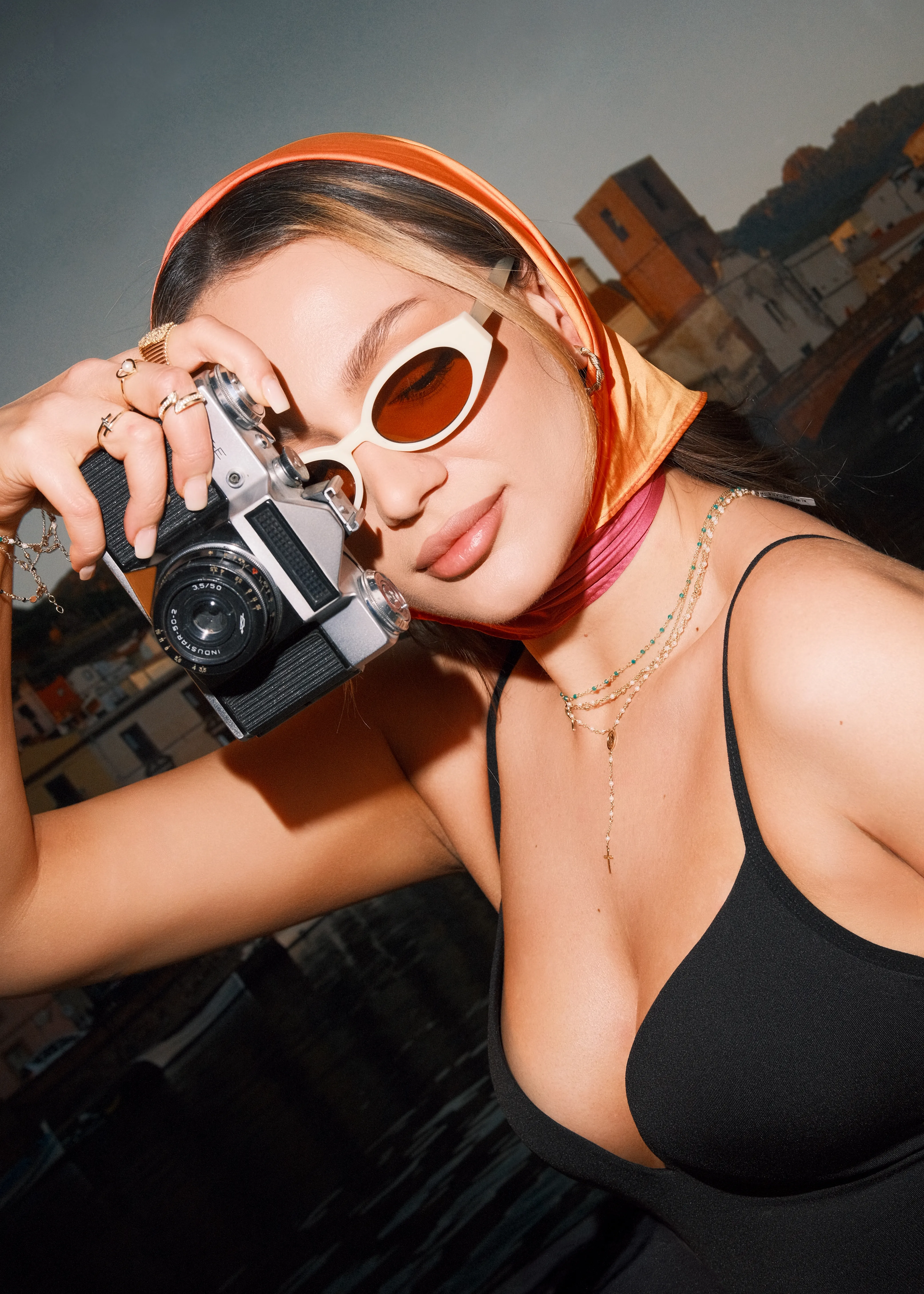 A model in an orange headscarf and sunglasses raises a vintage rangefinder camera to shoot, jewelry on her hand, a sunlit coastal town behind her