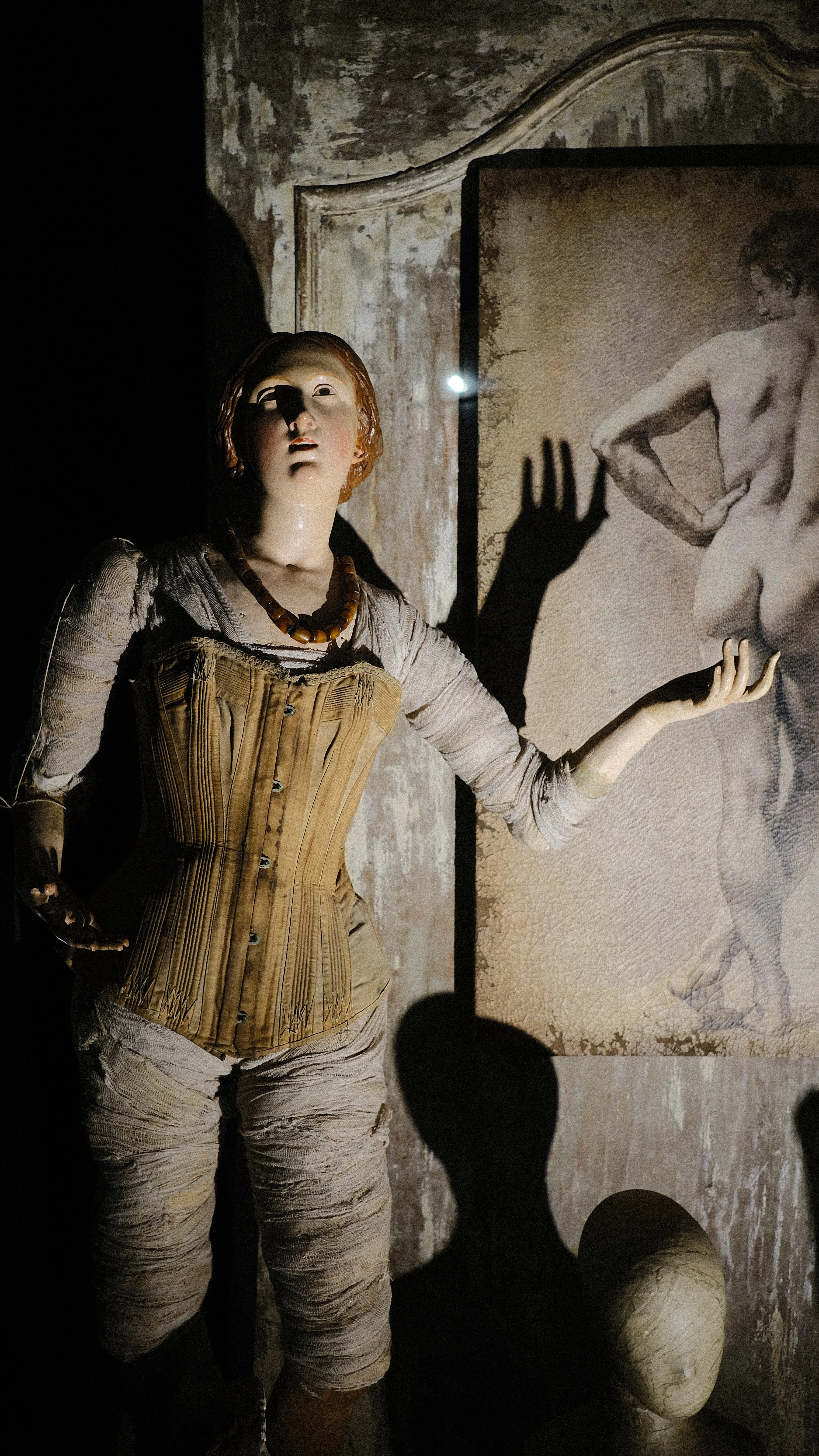 A weathered wax figure reaching toward a framed classical nude painting in dim theatrical light