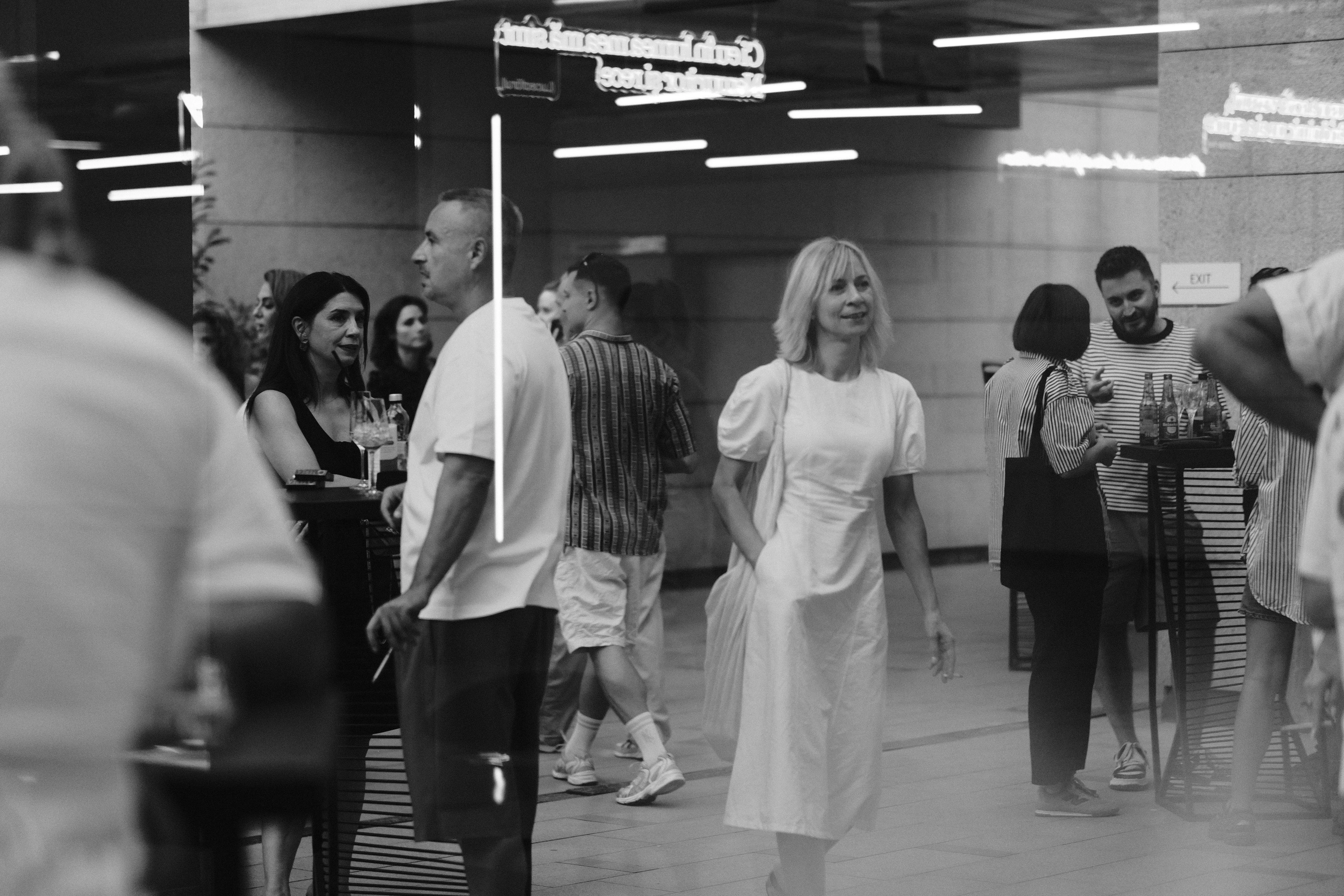 Guests in candid conversation reflected in glass beneath layered neon signage, black and white
