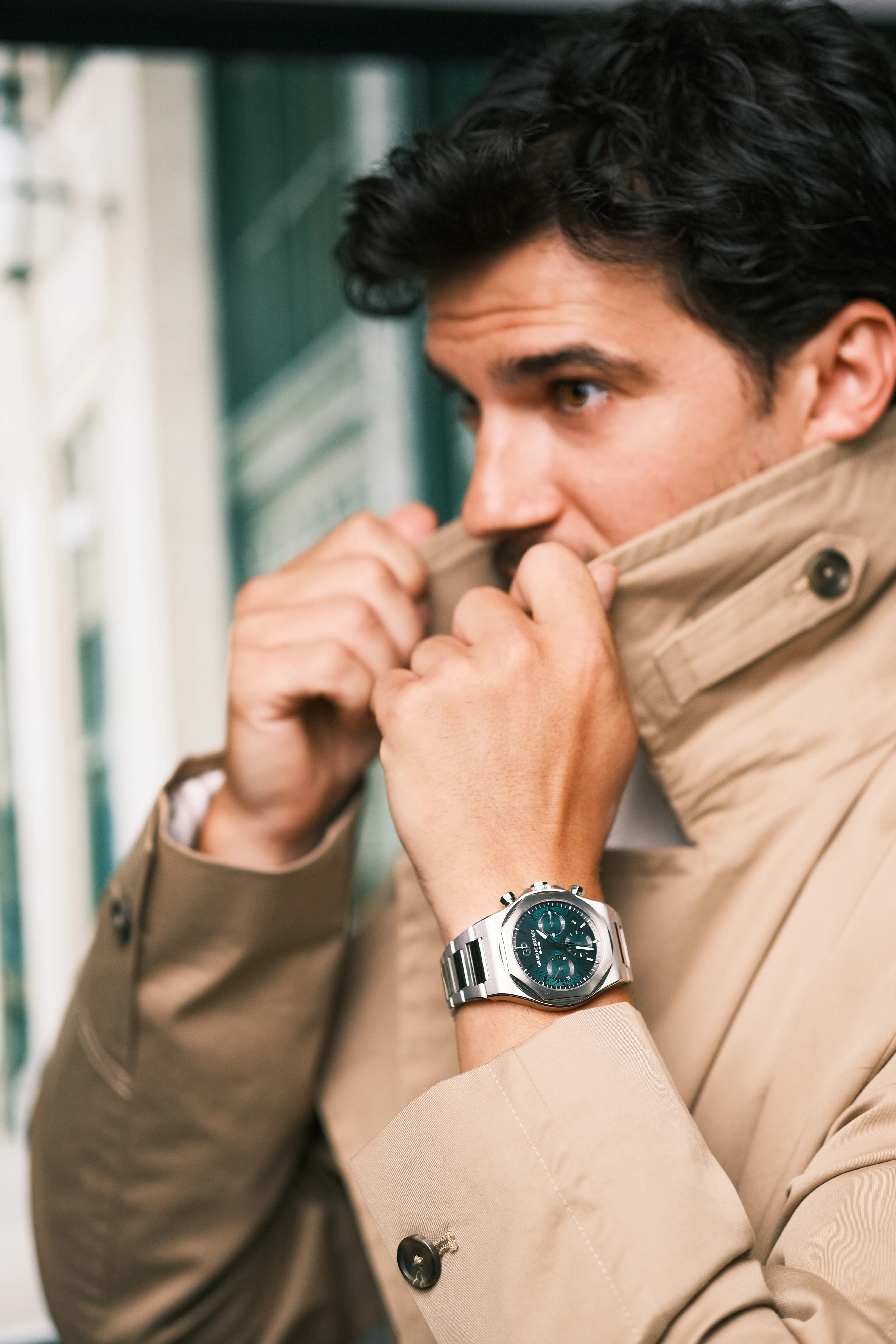 A man adjusts the collar of a trench coat, wristwatch visible, soft daylight