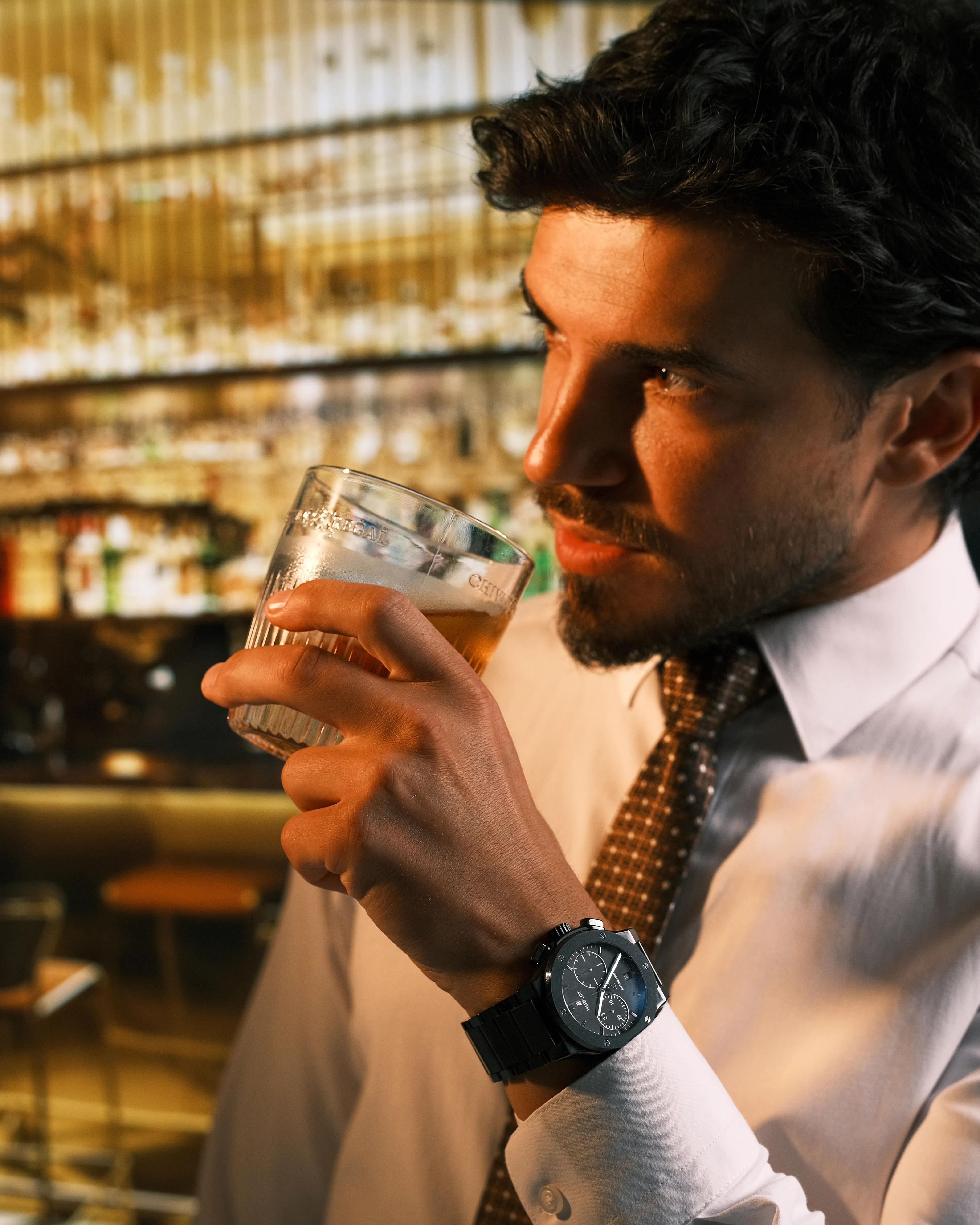 A man in a shirt and tie sips a drink at a bar, a chronograph watch visible on his wrist, warm bar lighting behind him