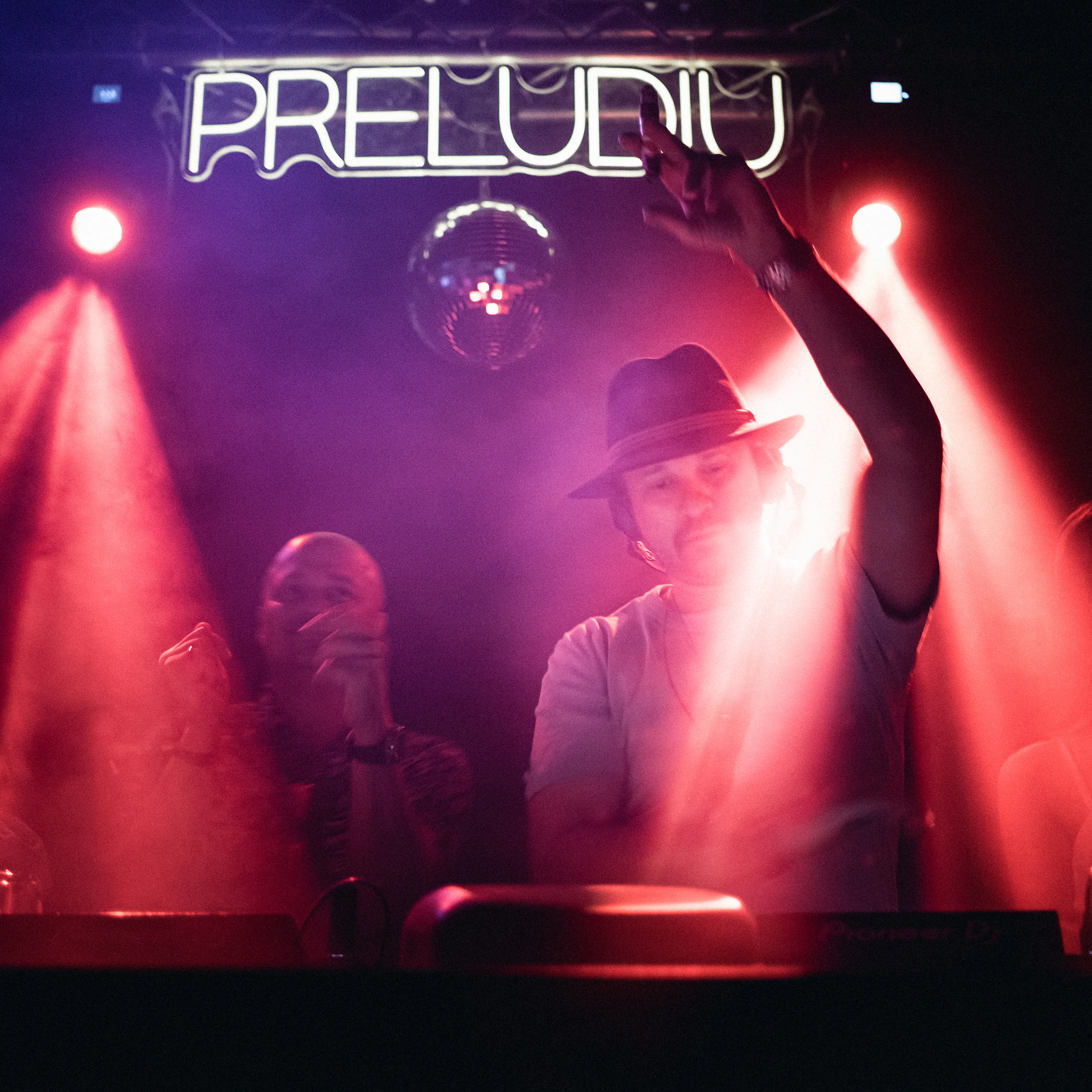 A DJ under a mirrorball beneath pink and purple stage light, one hand raised in the crowd