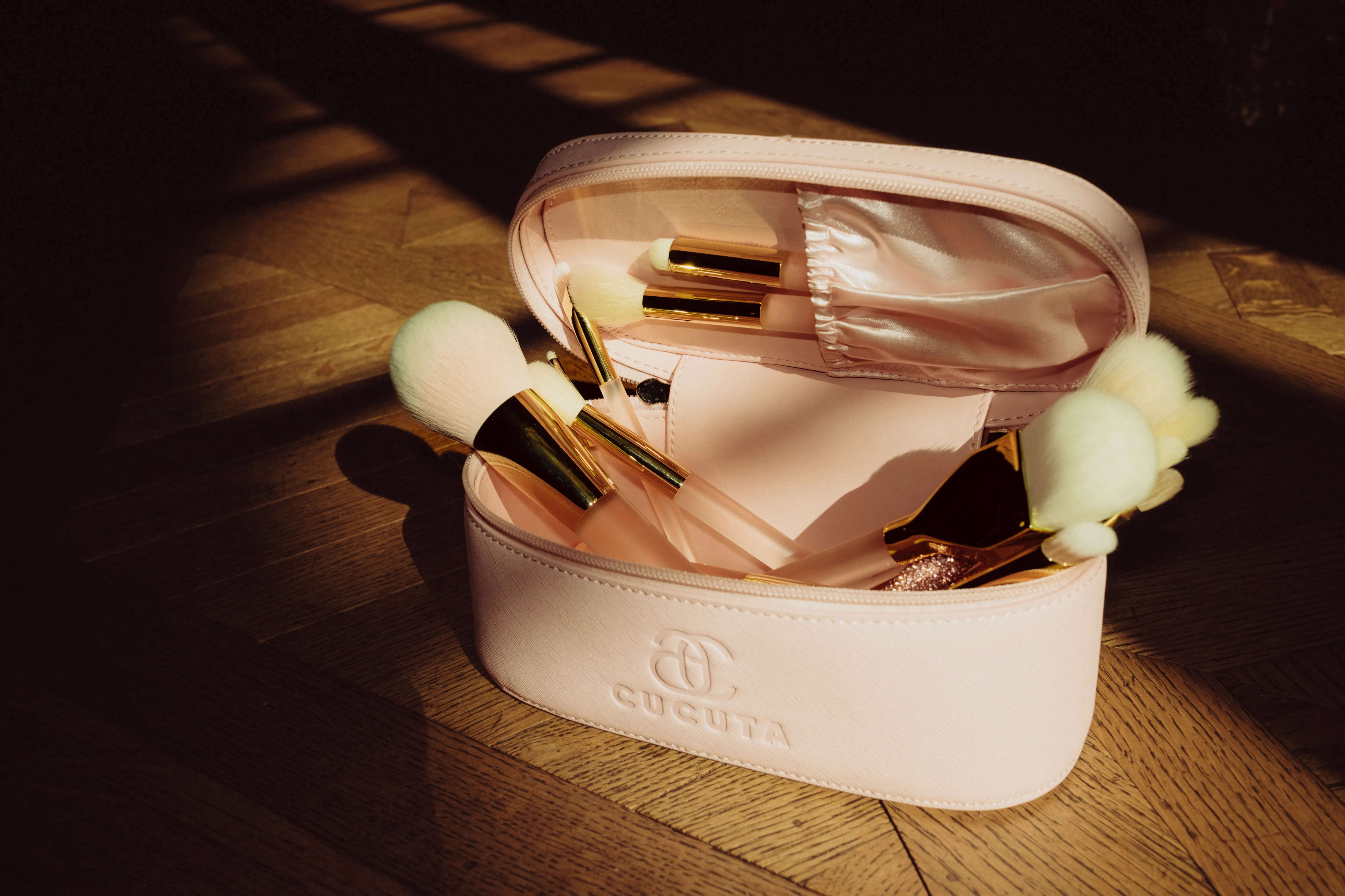 A pink cosmetic bag open with makeup brushes spilling out, dramatic sunlit shadow across a wood floor