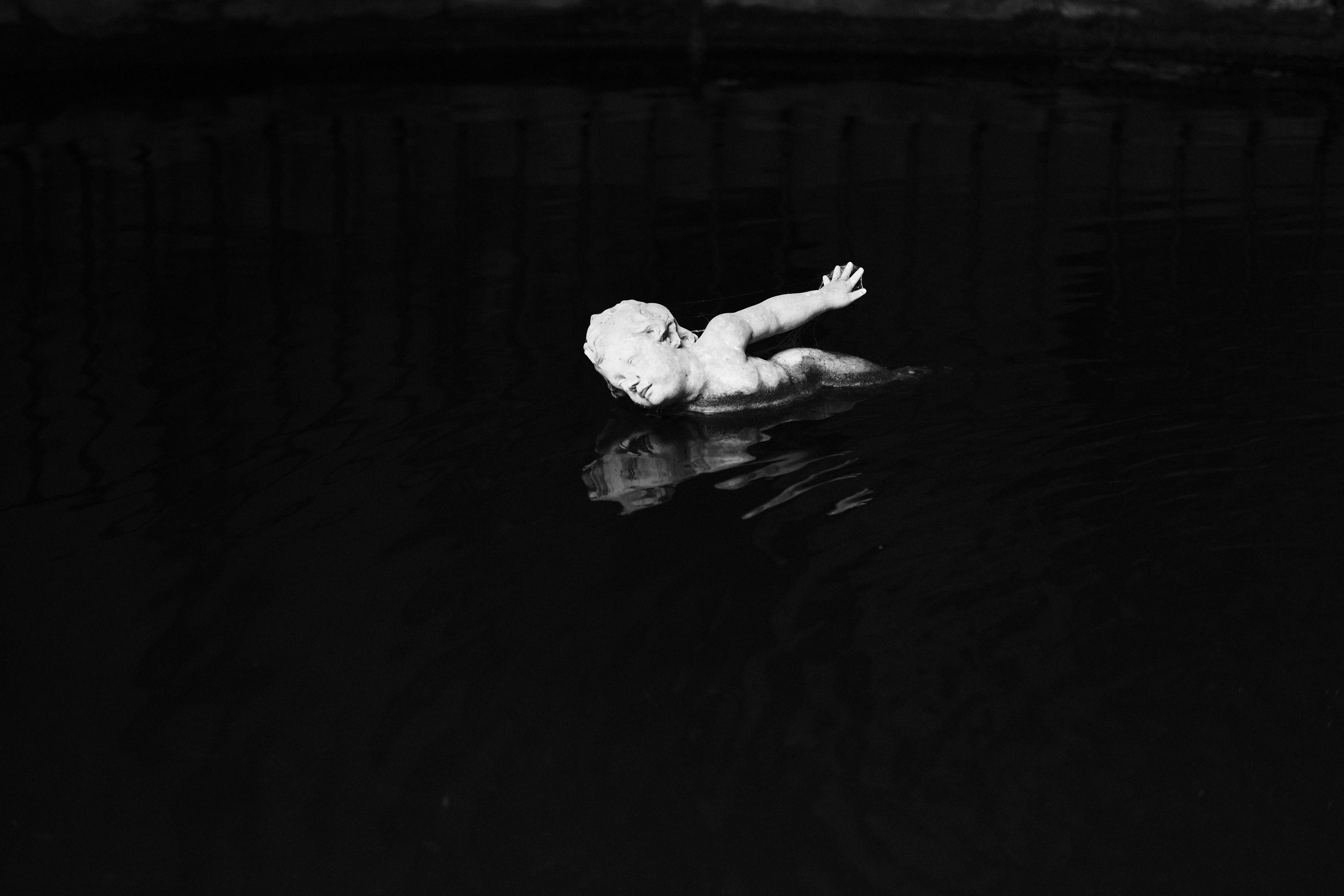 A marble cherub statue floating face-down in dark water, black and white