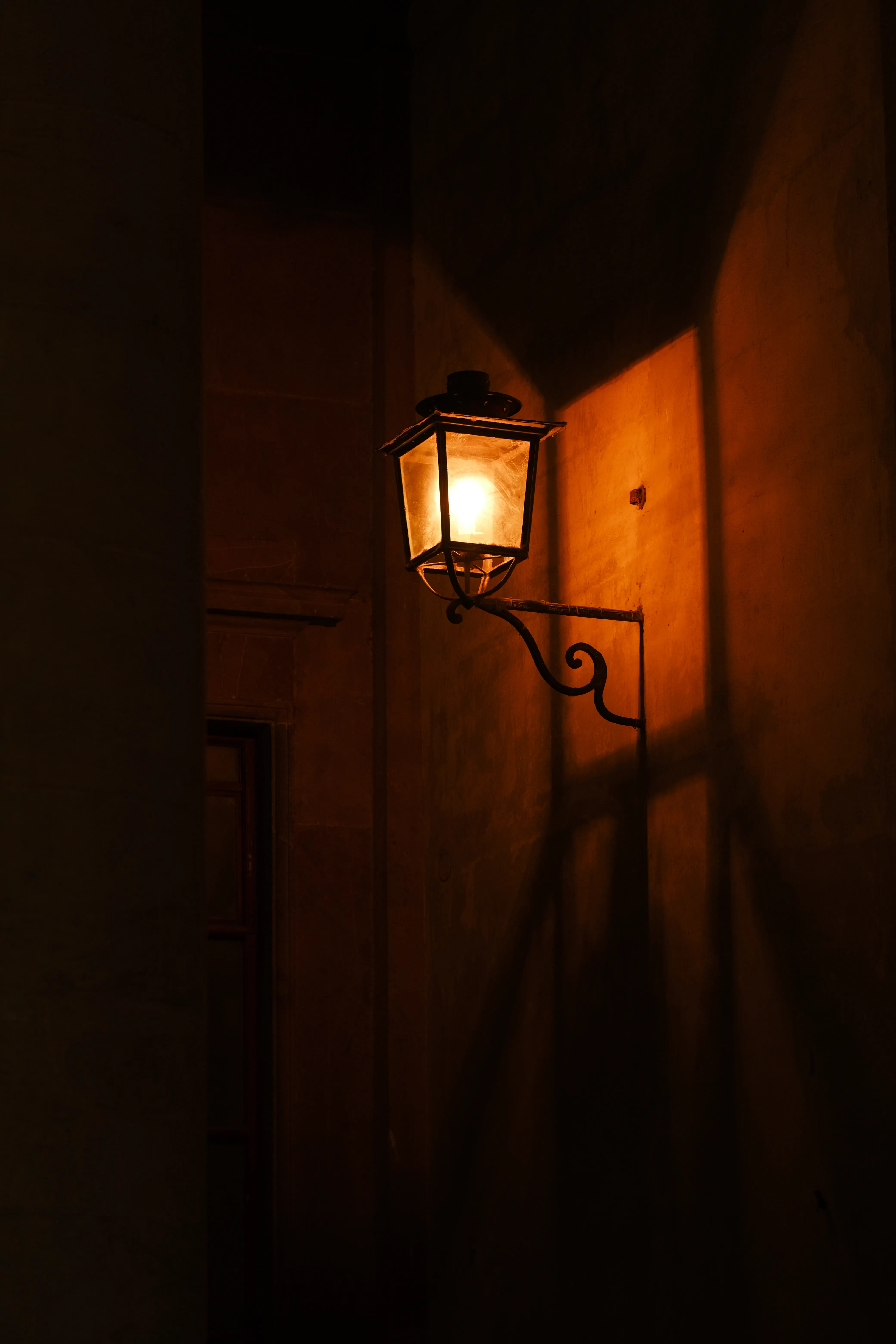 A single lit lantern glowing in a dark stone courtyard at night, warm light against deep shadow
