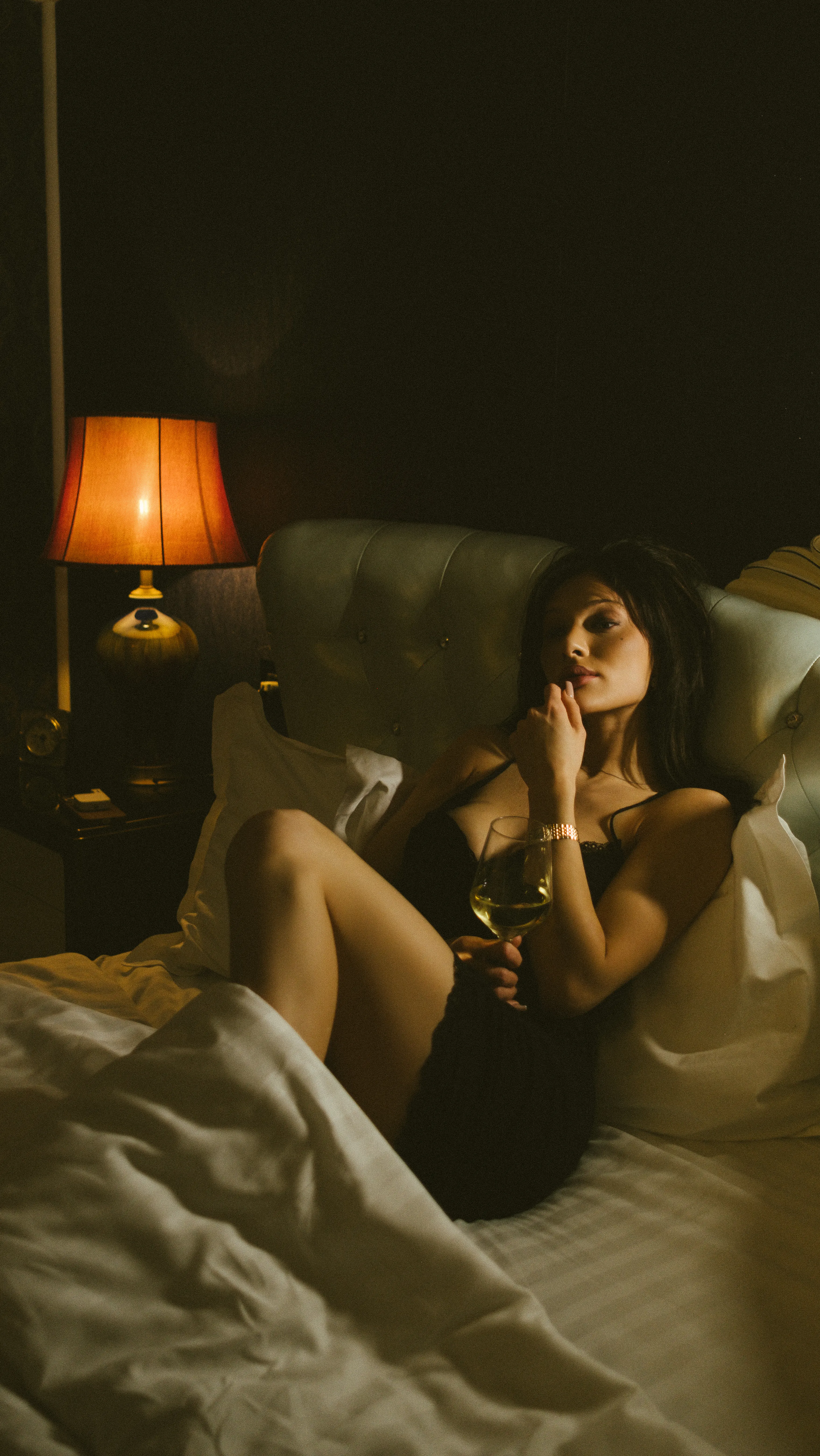 Warm, low-lit portrait in bed with a glass of wine, lamplight on one side