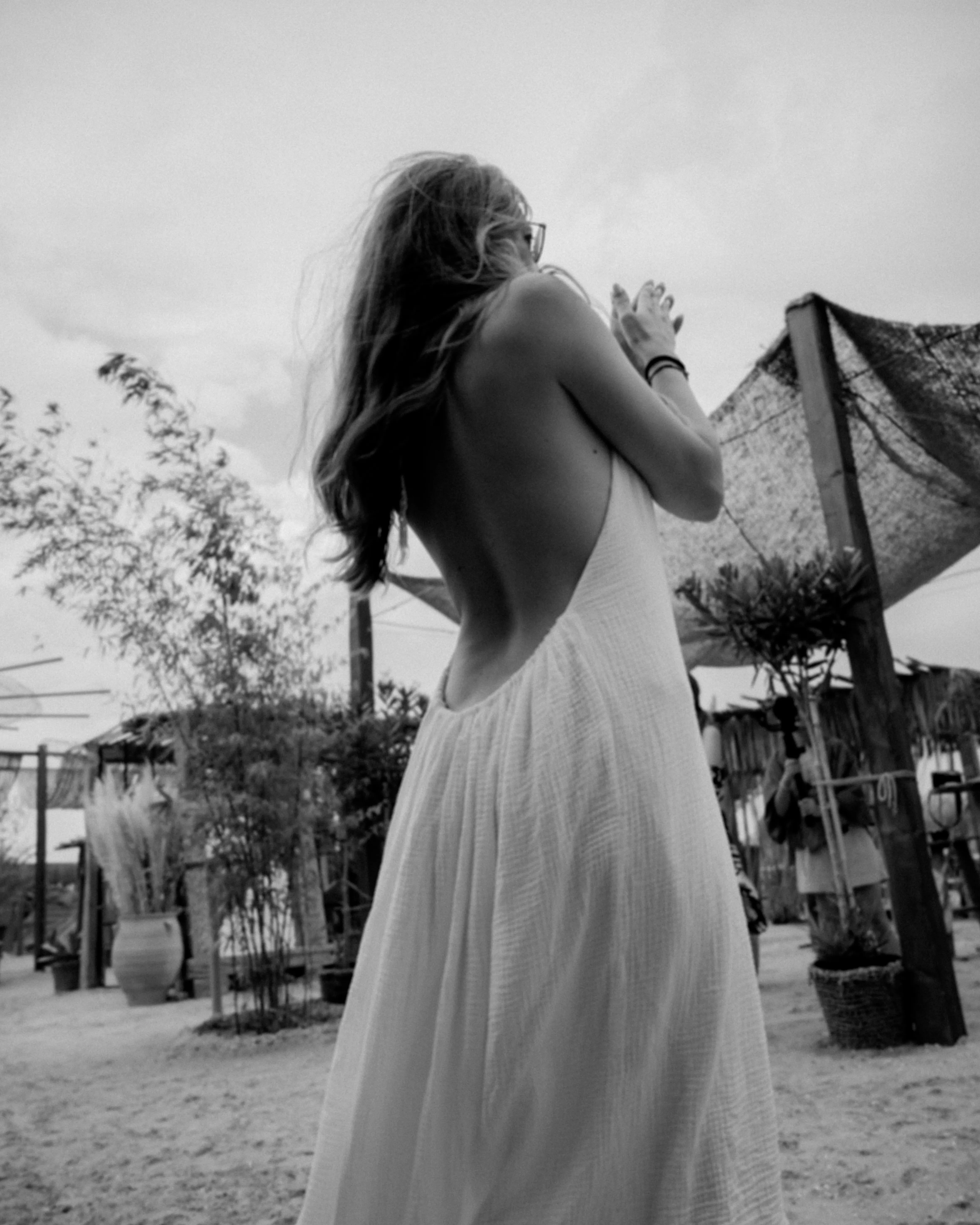 Black and white photo of a woman in a backless dress, hair blown by wind, standing before a thatched beach-club structure