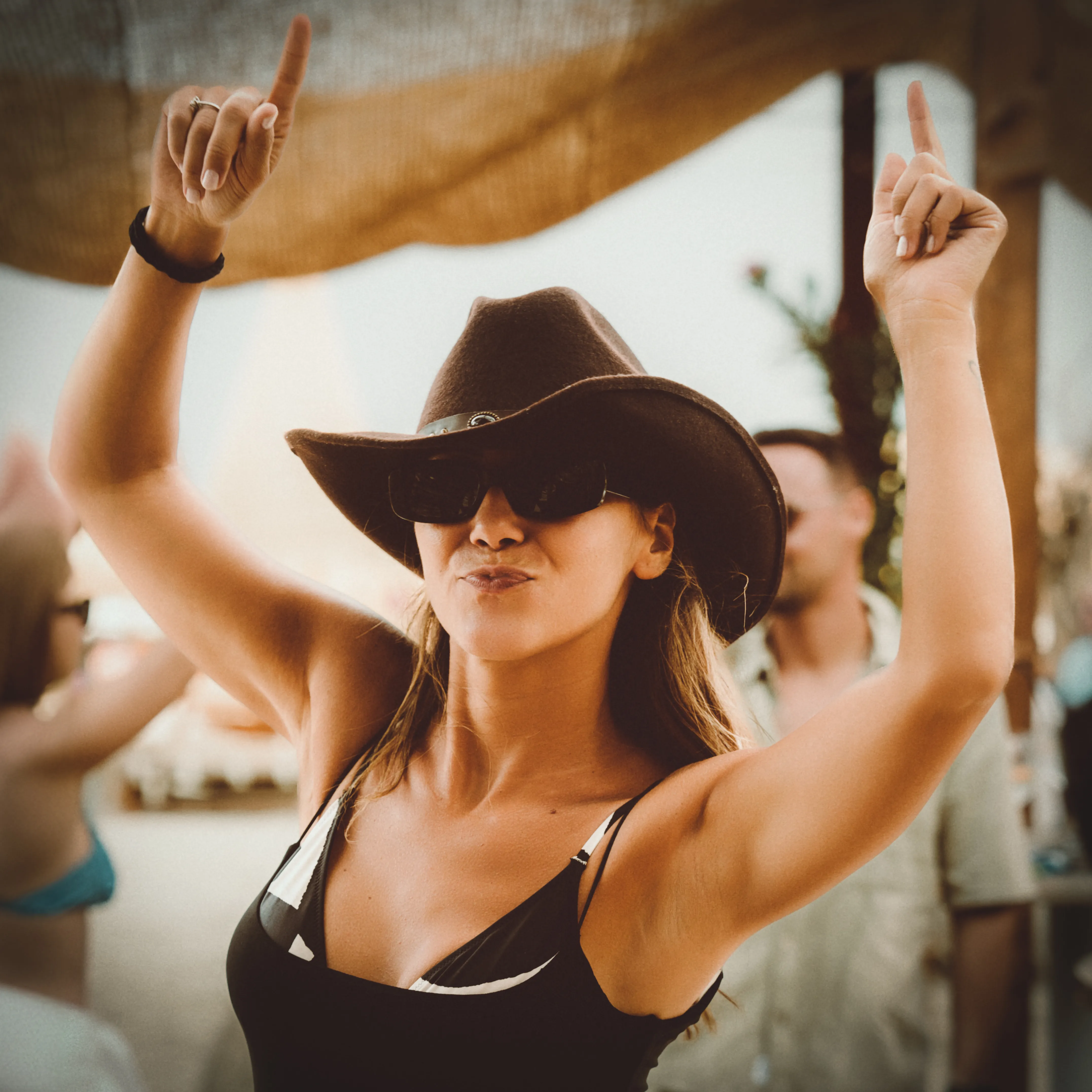 A woman in a wide-brimmed hat and sunglasses dances with both arms raised under a canopy at golden hour
