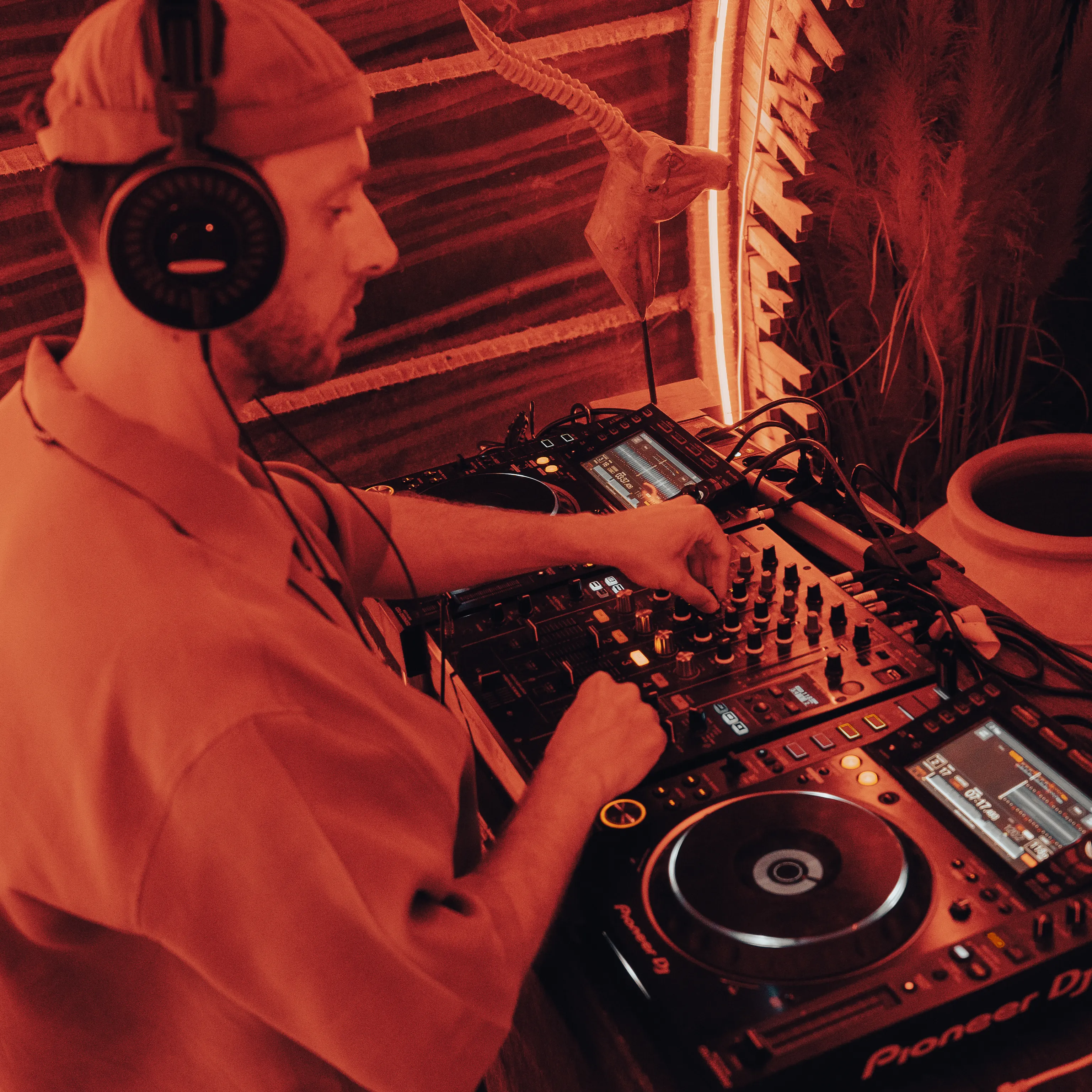 A DJ adjusts a mixer under red light, headphones on, in a thatched-roof booth