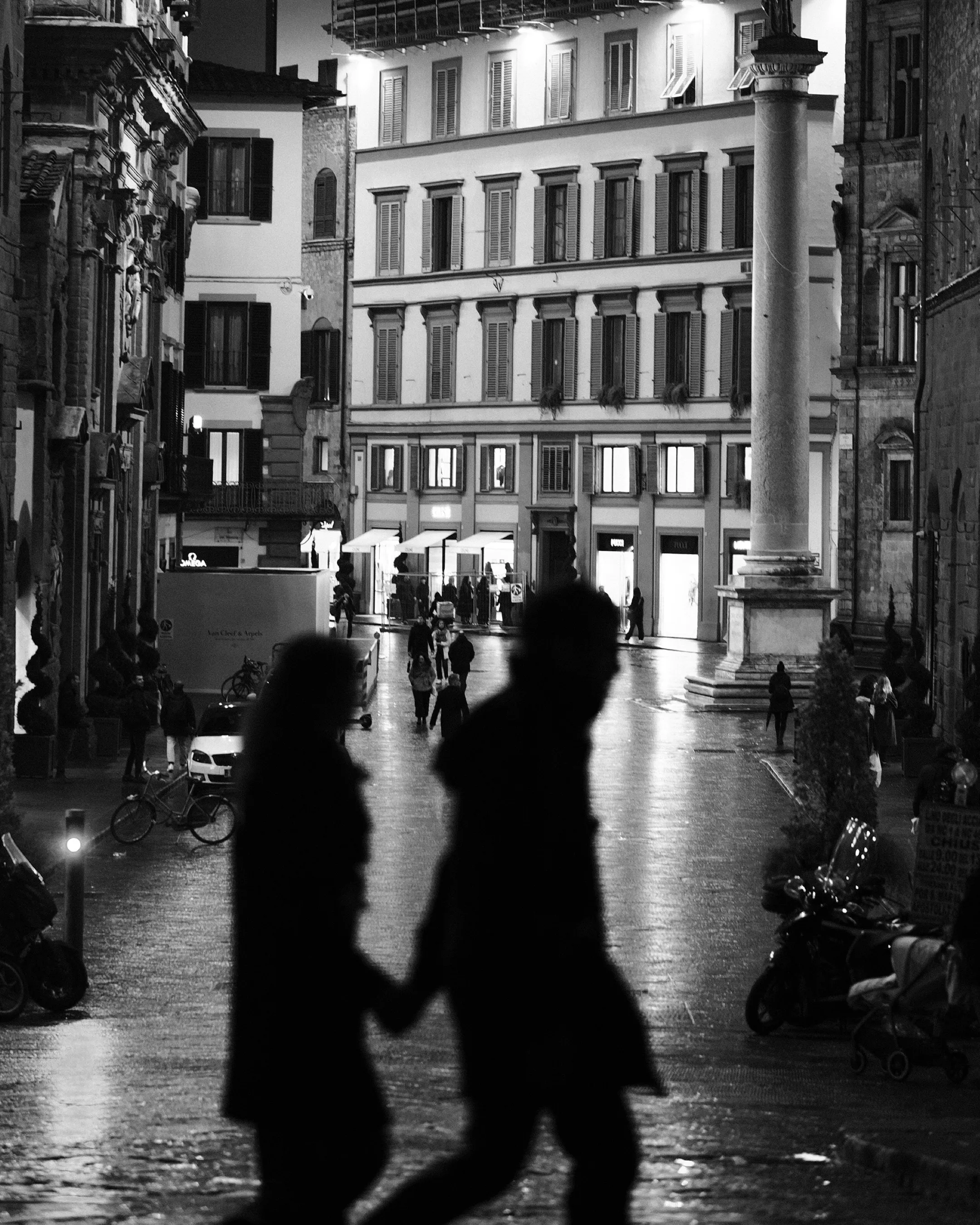 Two silhouetted figures hold hands crossing a piazza at night, Florentine architecture lit behind them