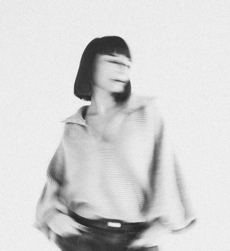 Motion-blurred black and white portrait — hair swept across the face, hands in the pockets of a ribbed sweater, grain and blur intentional