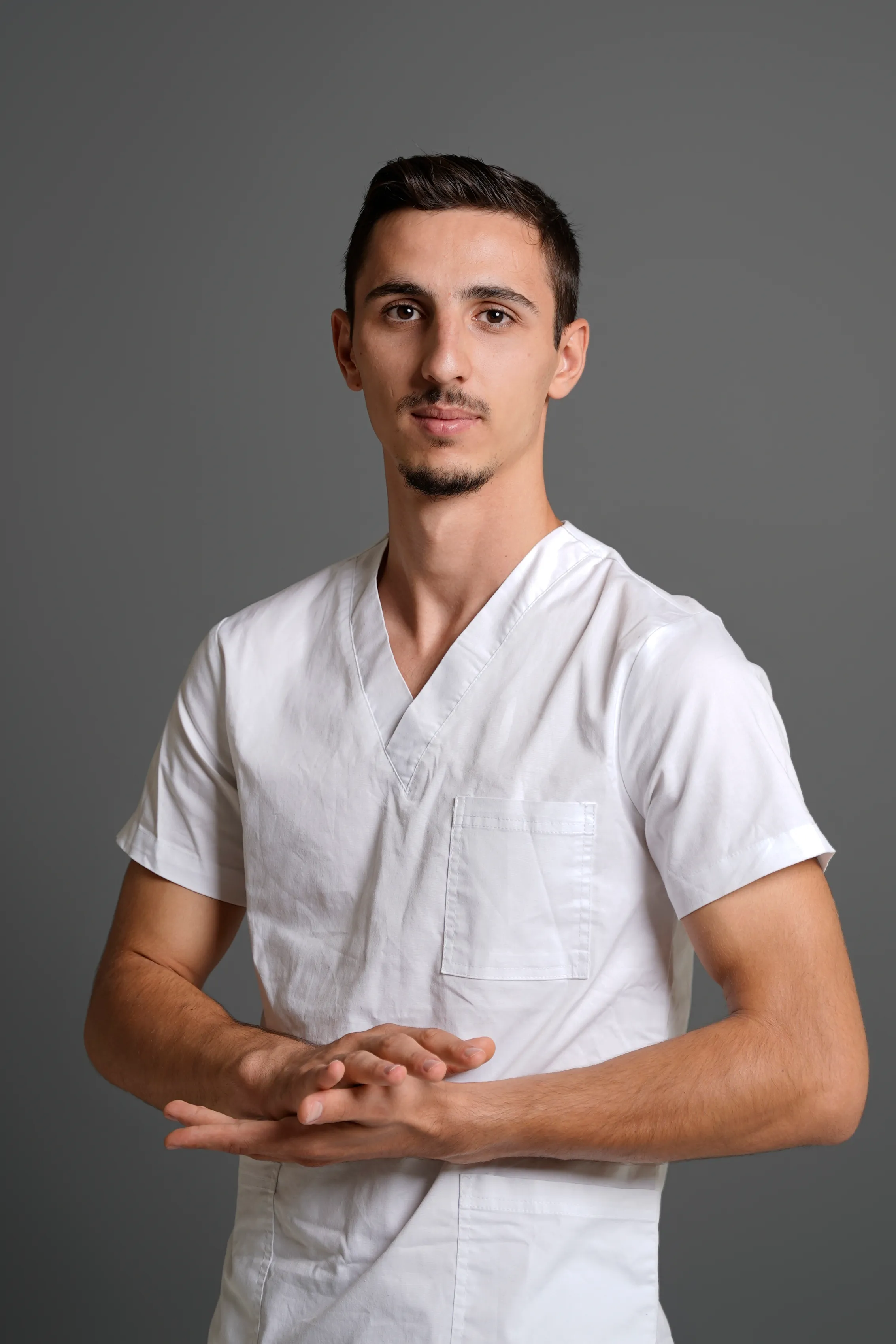 Man in white medical scrubs, hands clasped, grey studio background