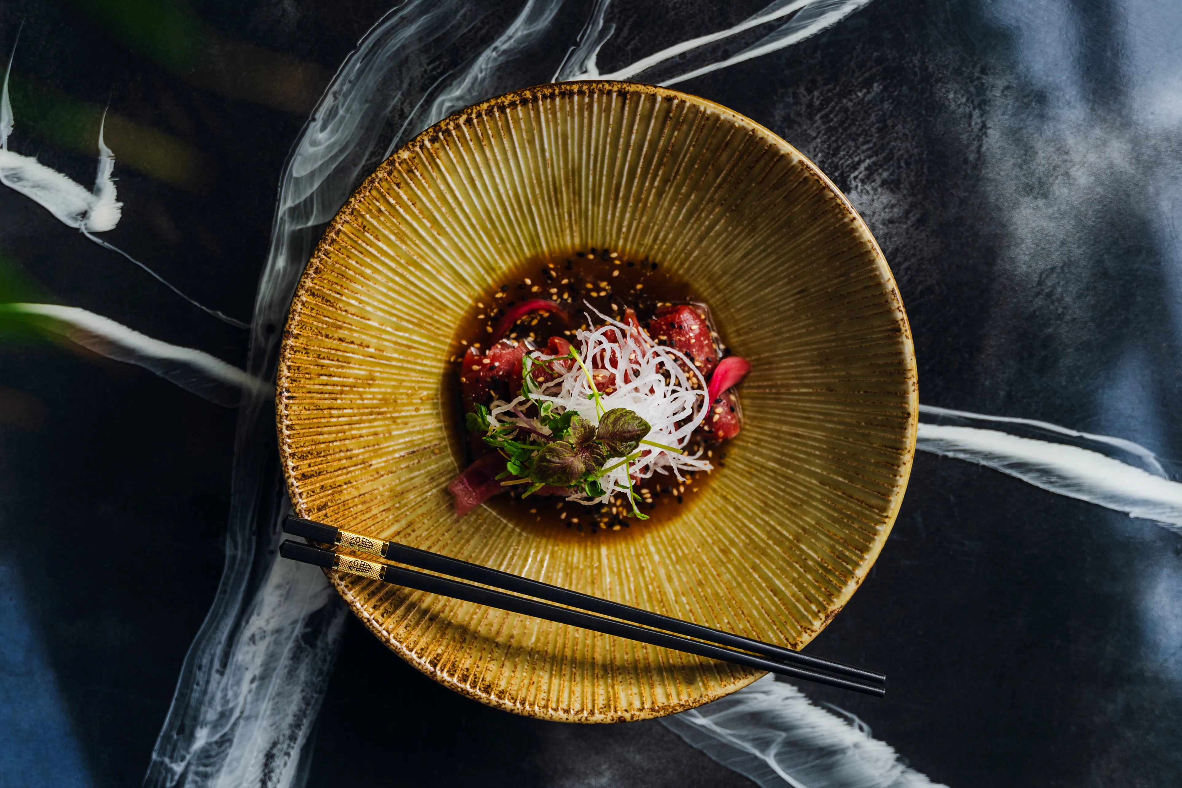 An overhead shot of a tuna dish in dark broth, plated on a ridged ceramic bowl with black chopsticks