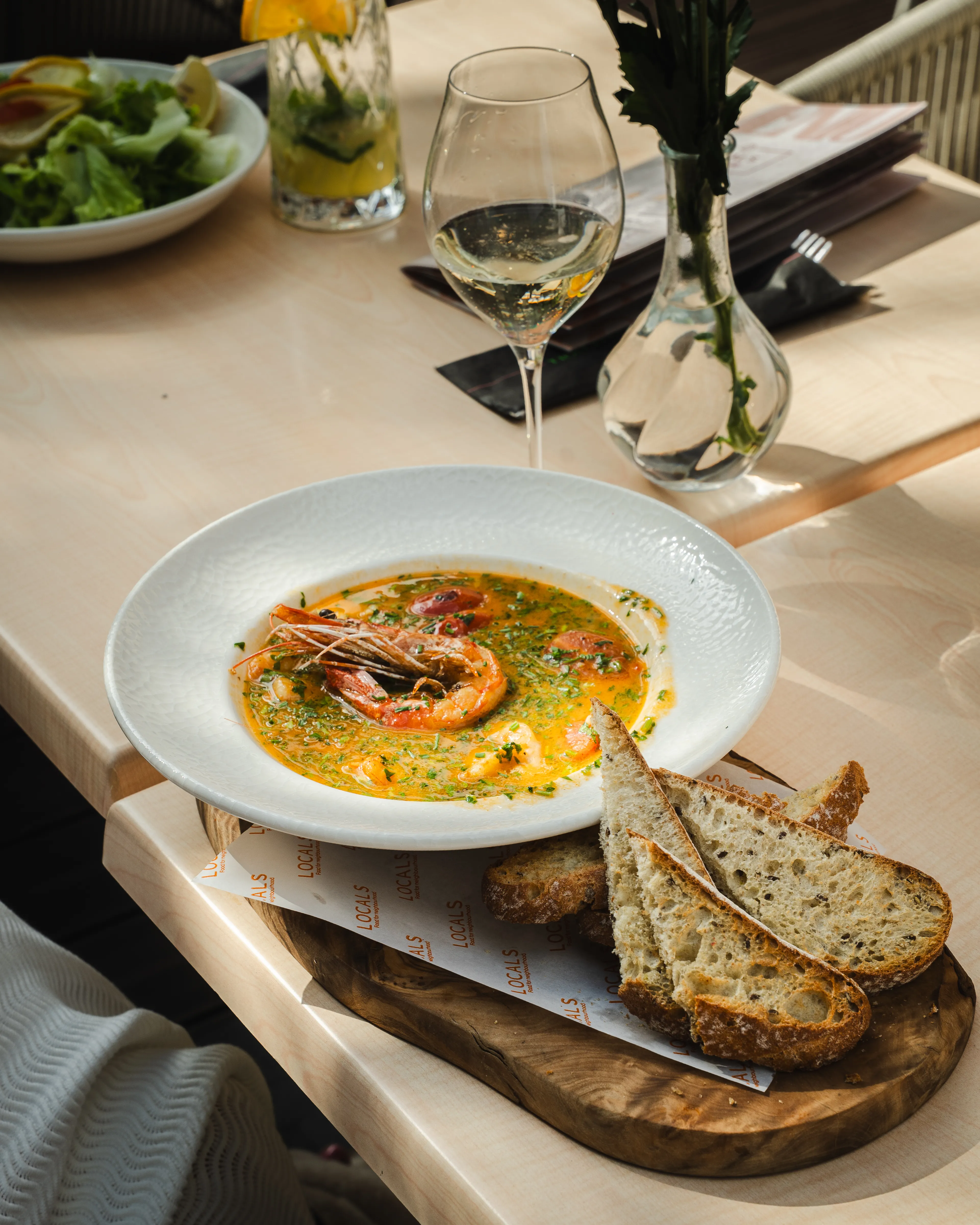 A sunlit restaurant table with a seafood soup, bread, a glass of wine, and a spritz