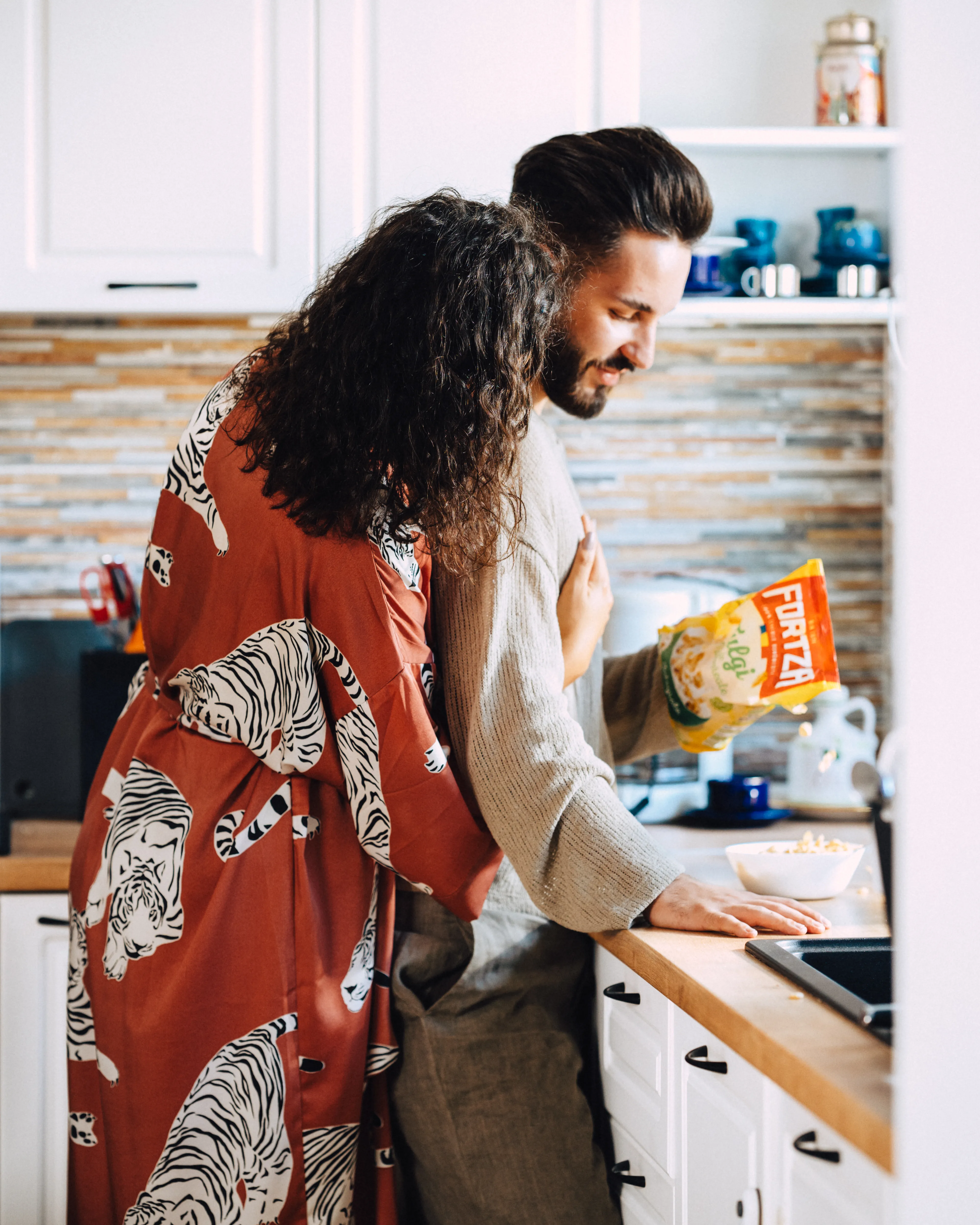 A couple laughing together in a home kitchen, cereal box on the counter between them