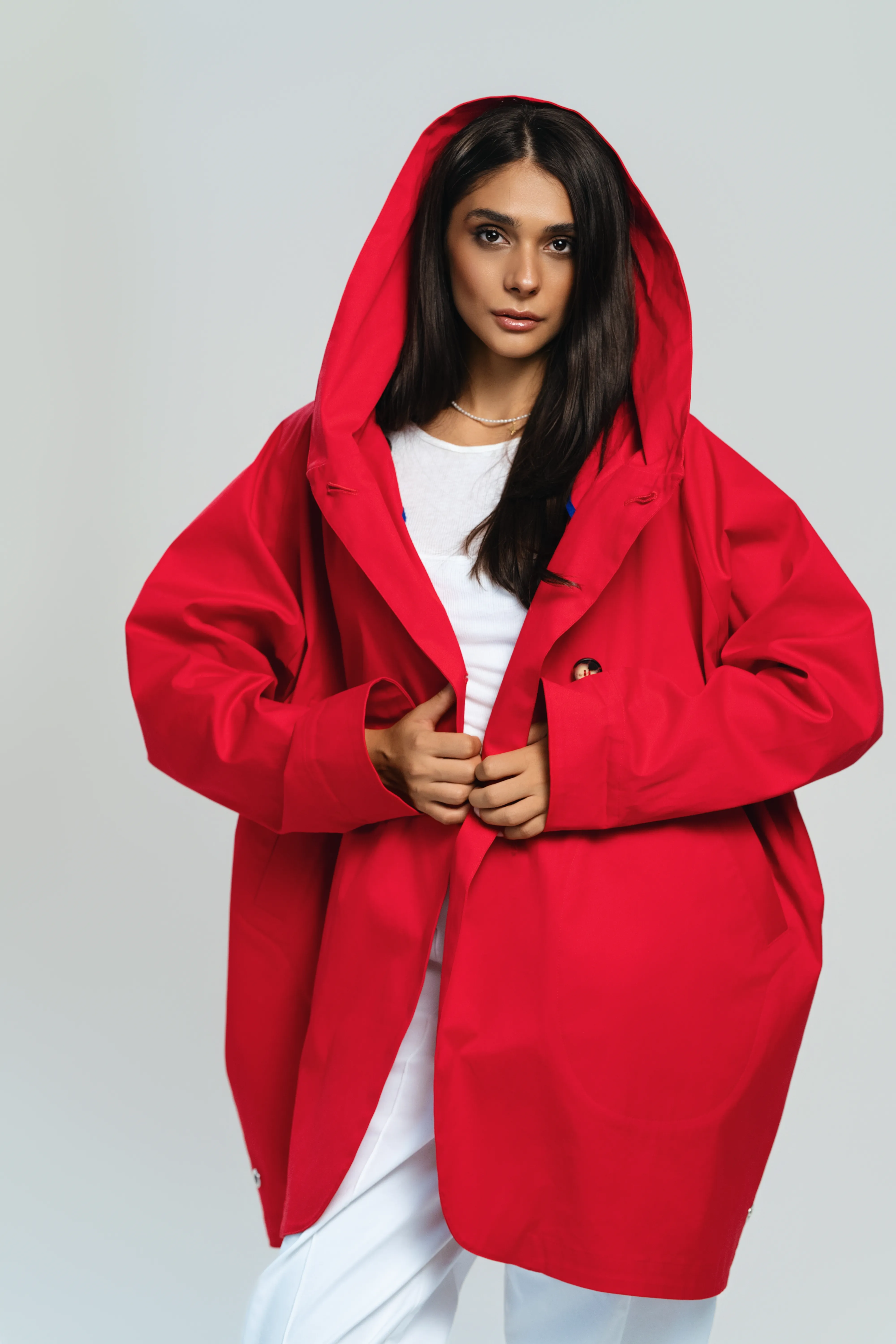 A model in an oversized red hooded coat looks directly at camera, hands clasped at the lapel, studio grey background