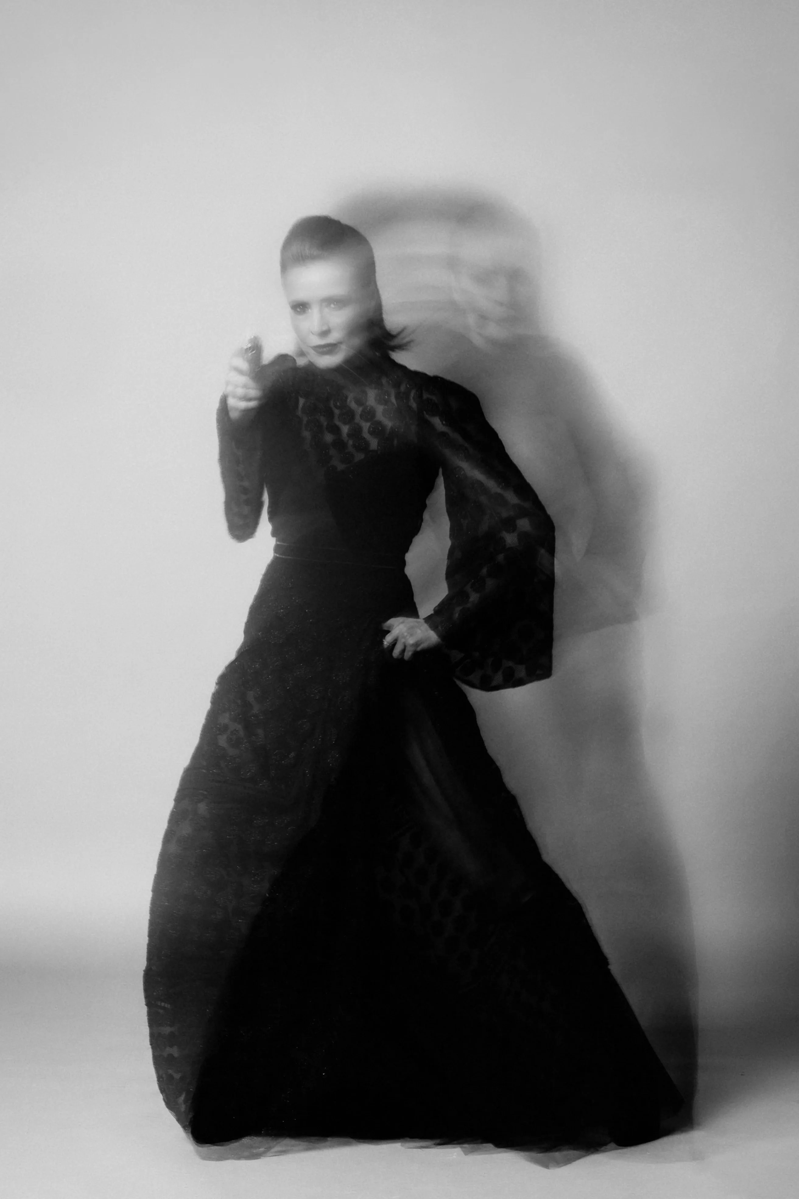 A figure in an ornate lace gown, doubled by motion blur, gesturing toward the camera