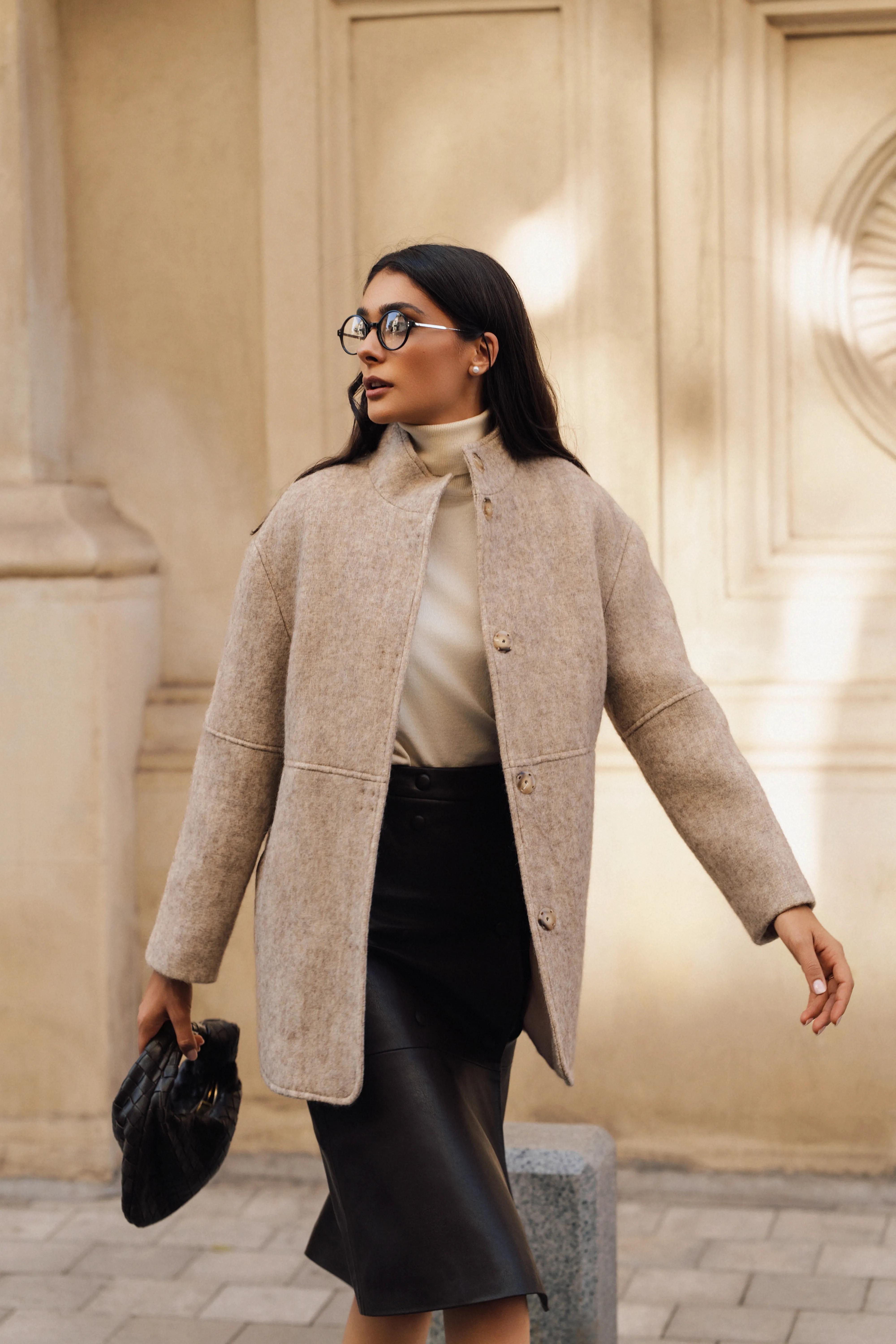 Model in a camel wool coat and black skirt walking past a colonnade, sunglasses