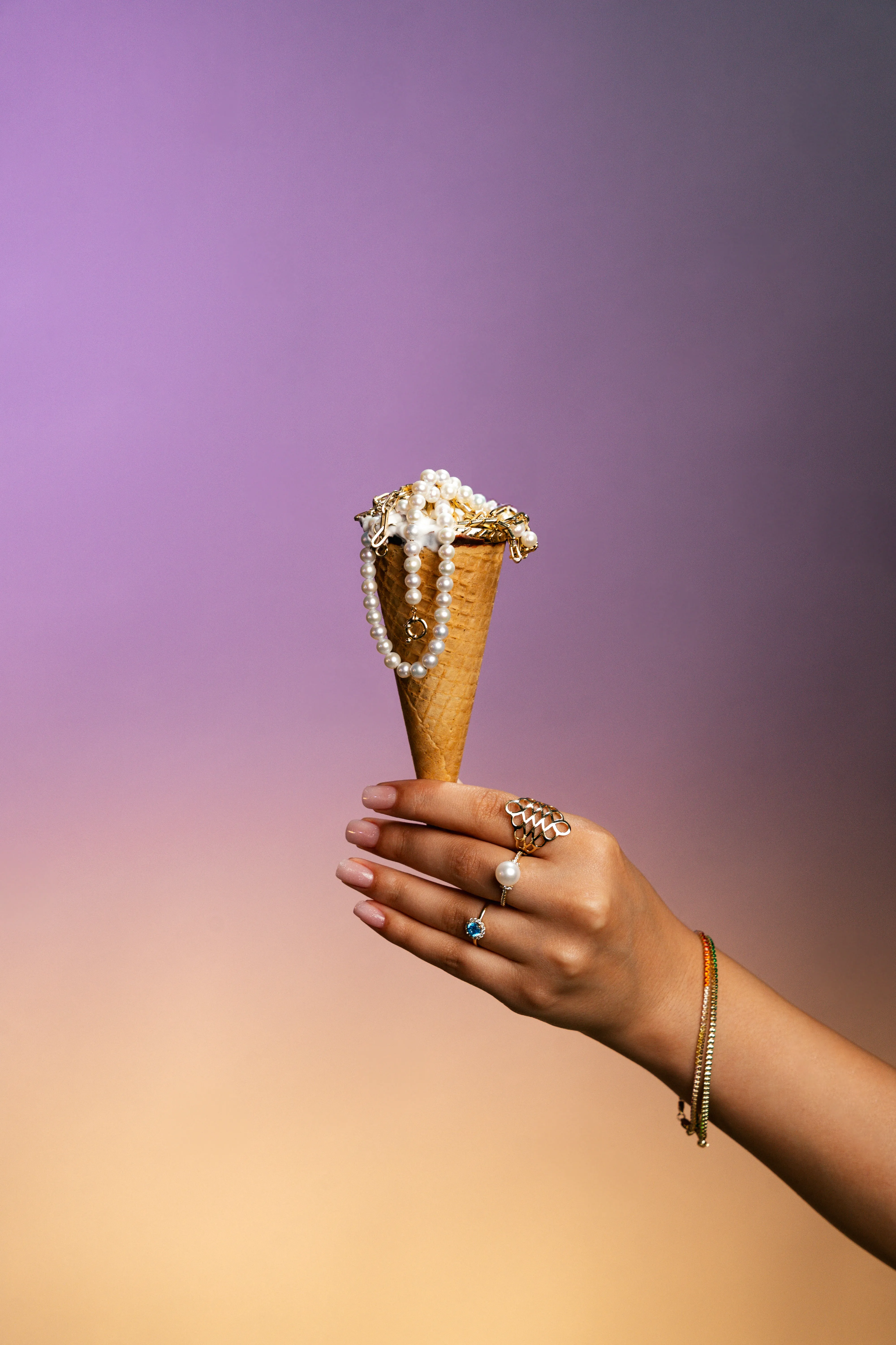 An ice cream cone dripping with pearls and gold jewelry instead of ice cream, on a purple-to-peach gradient
