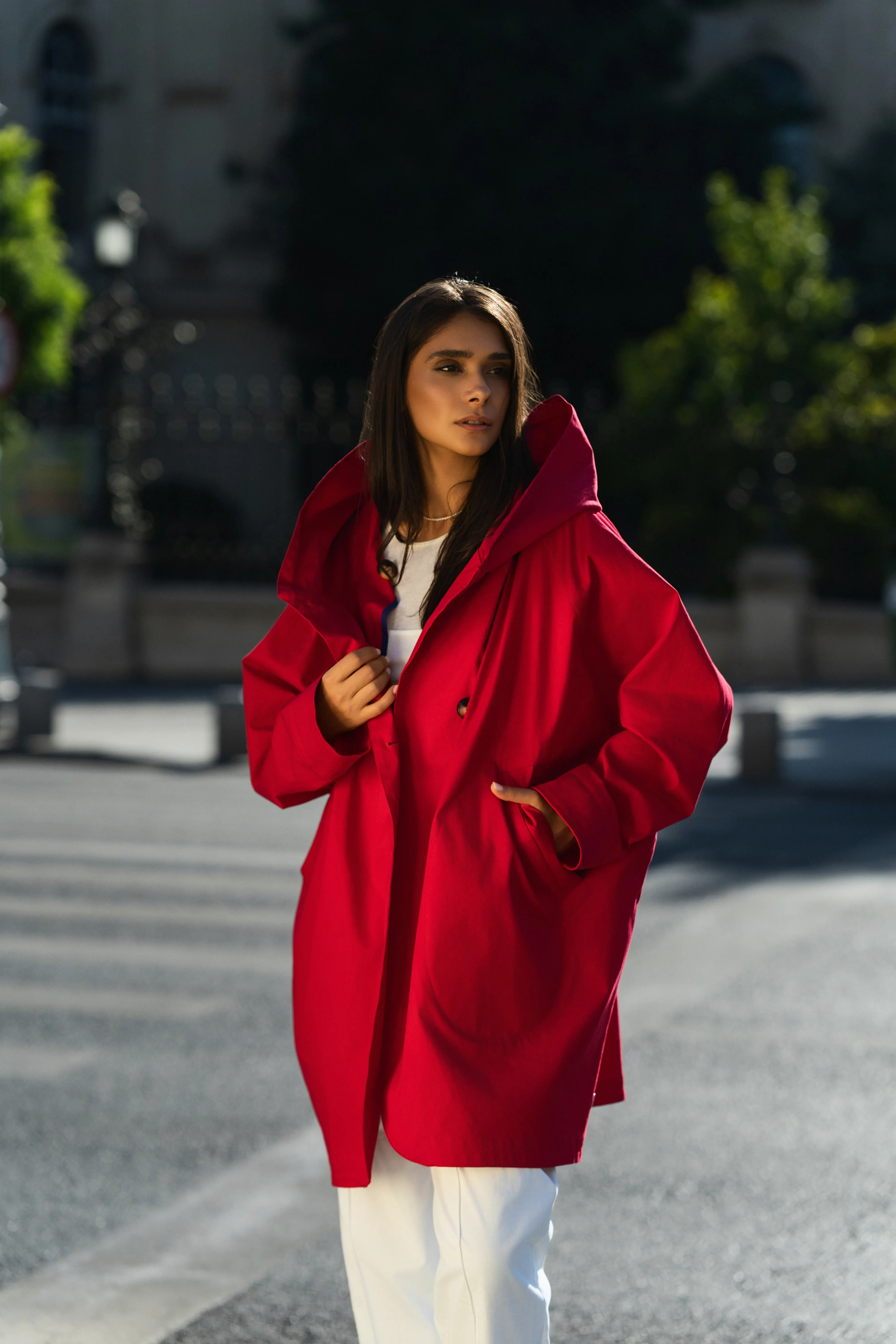 Model in an oversized red hooded coat on a city street in golden side-light, hand in pocket